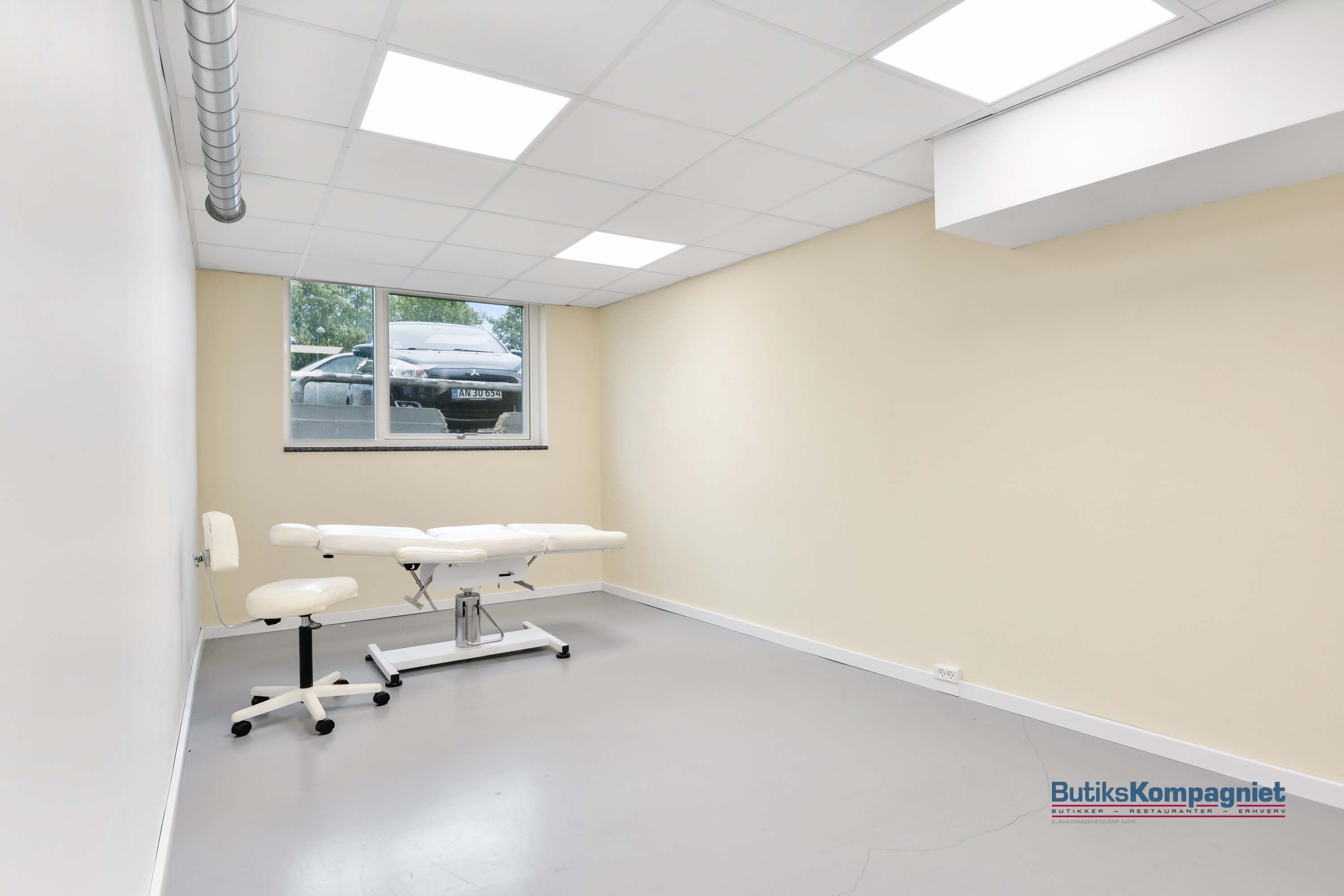 En tom stue med en hvid massagebord, en hvid stol og lyse loftslampe. Der er et vindue med udsigt til en parkeringsplads med