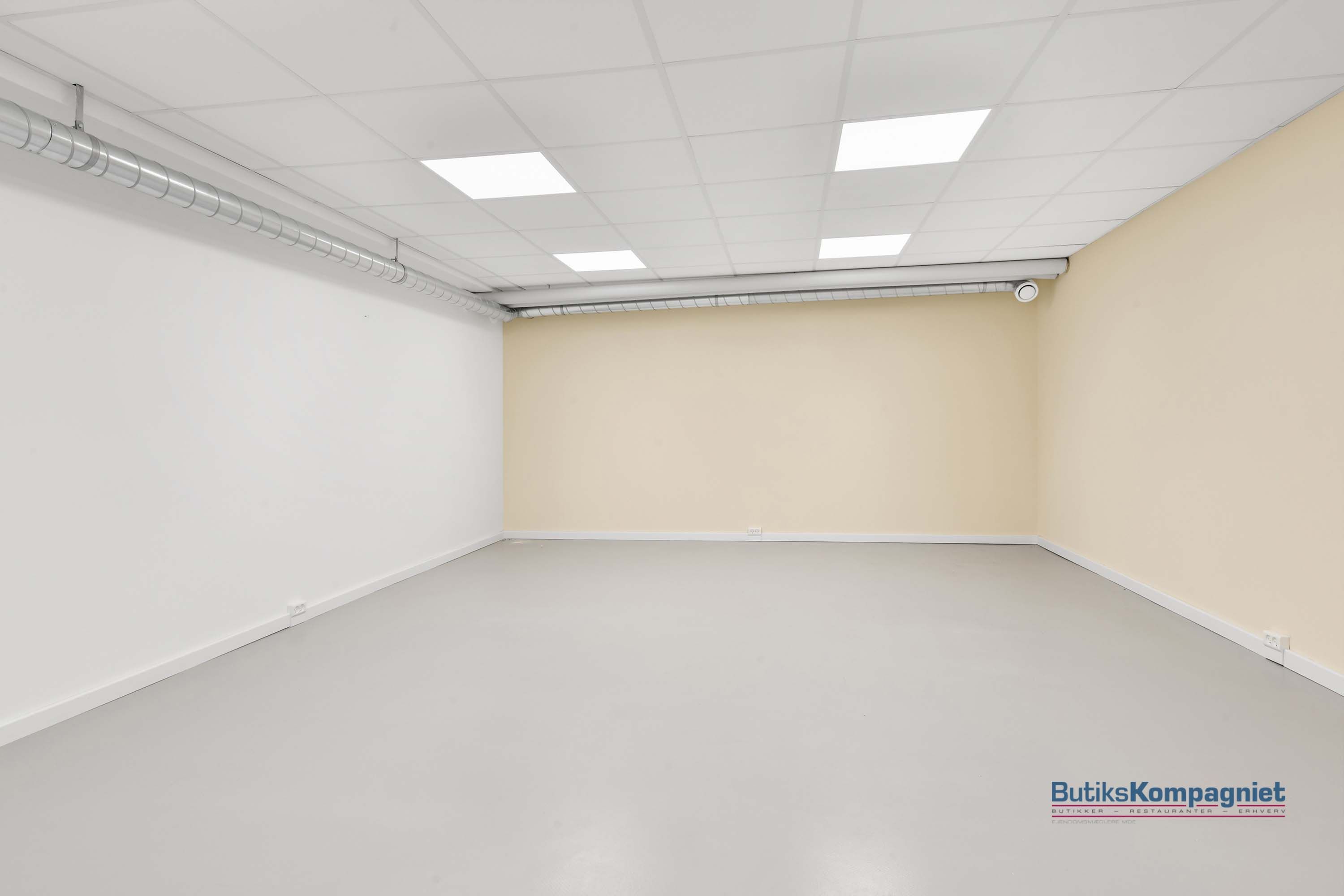 Rum med hvidt loft og gulv, samt en lys beige væg. Rundt om rummet er der elektriske ledninger og to hvide elektriske stik.