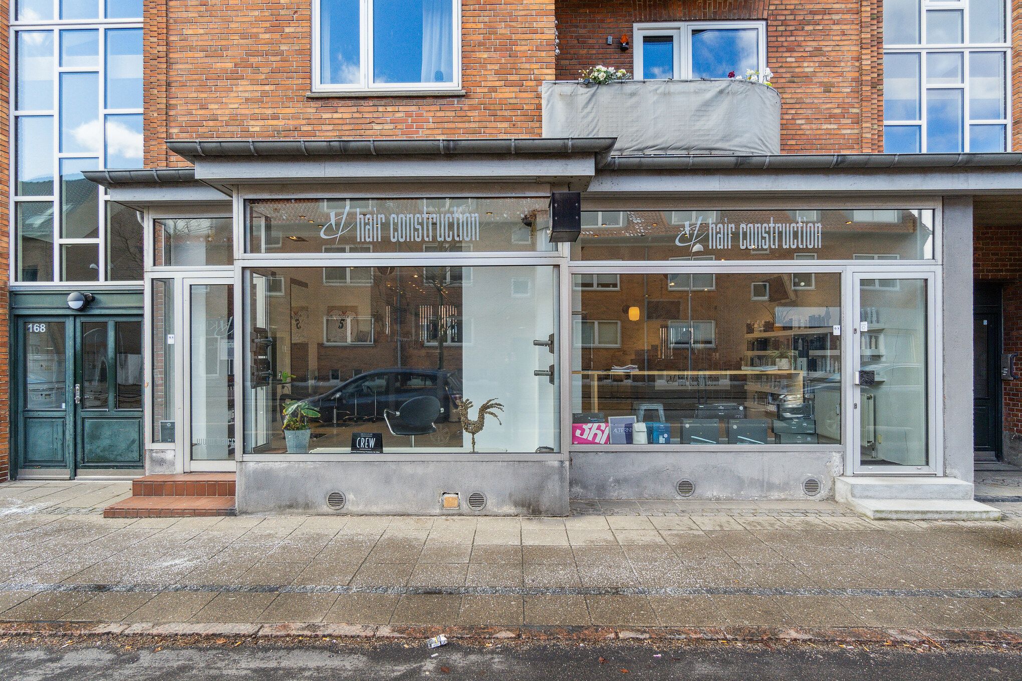 Fremtrædende facade med glassdøre og store vinduer siger Hair Construction. Inde er der en række stole og et gulbord med