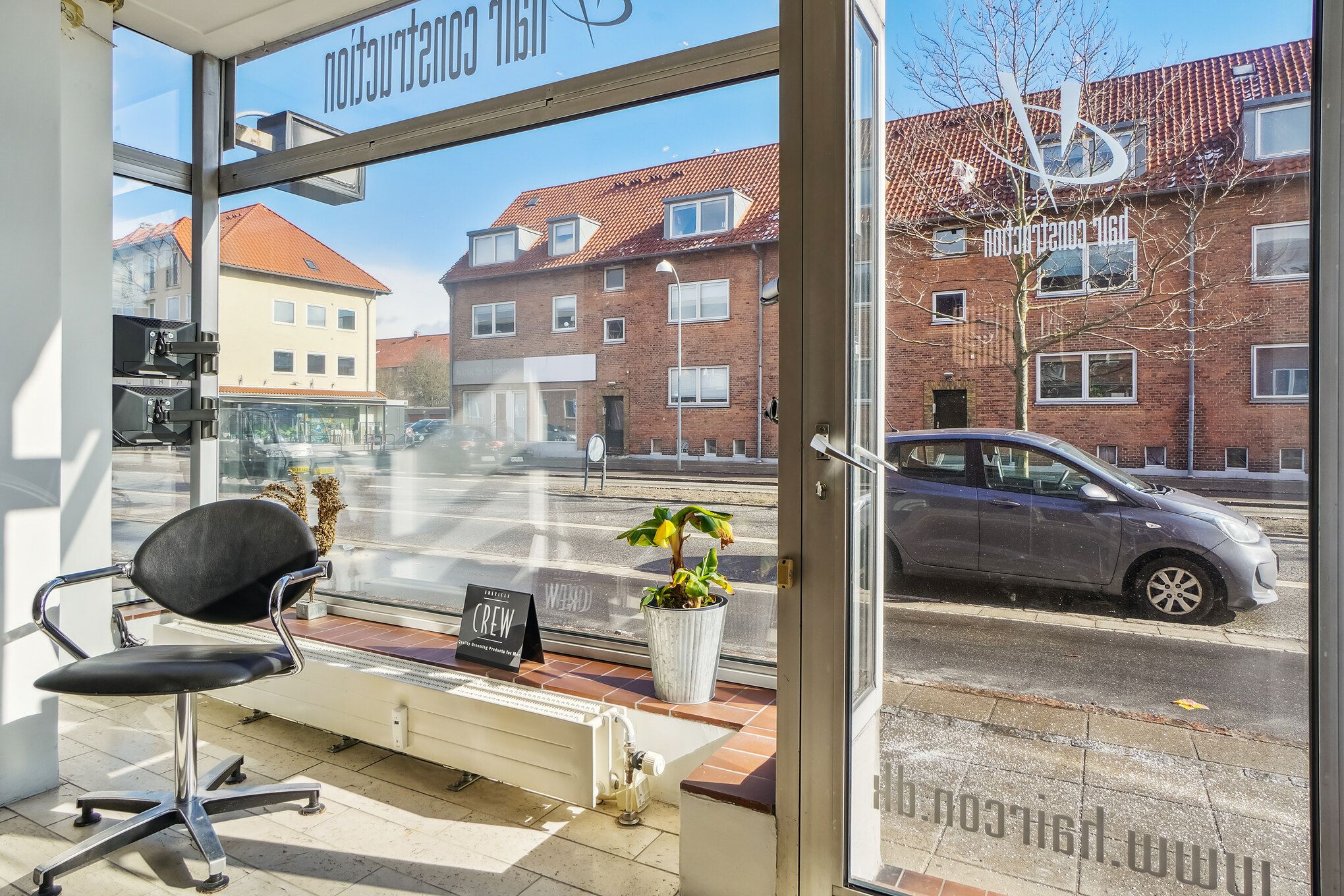 Oplysninger om frisørsalonen American Crew med en kontormøbel og en plante på vindueskarmen med udsigt til en parkeret bil