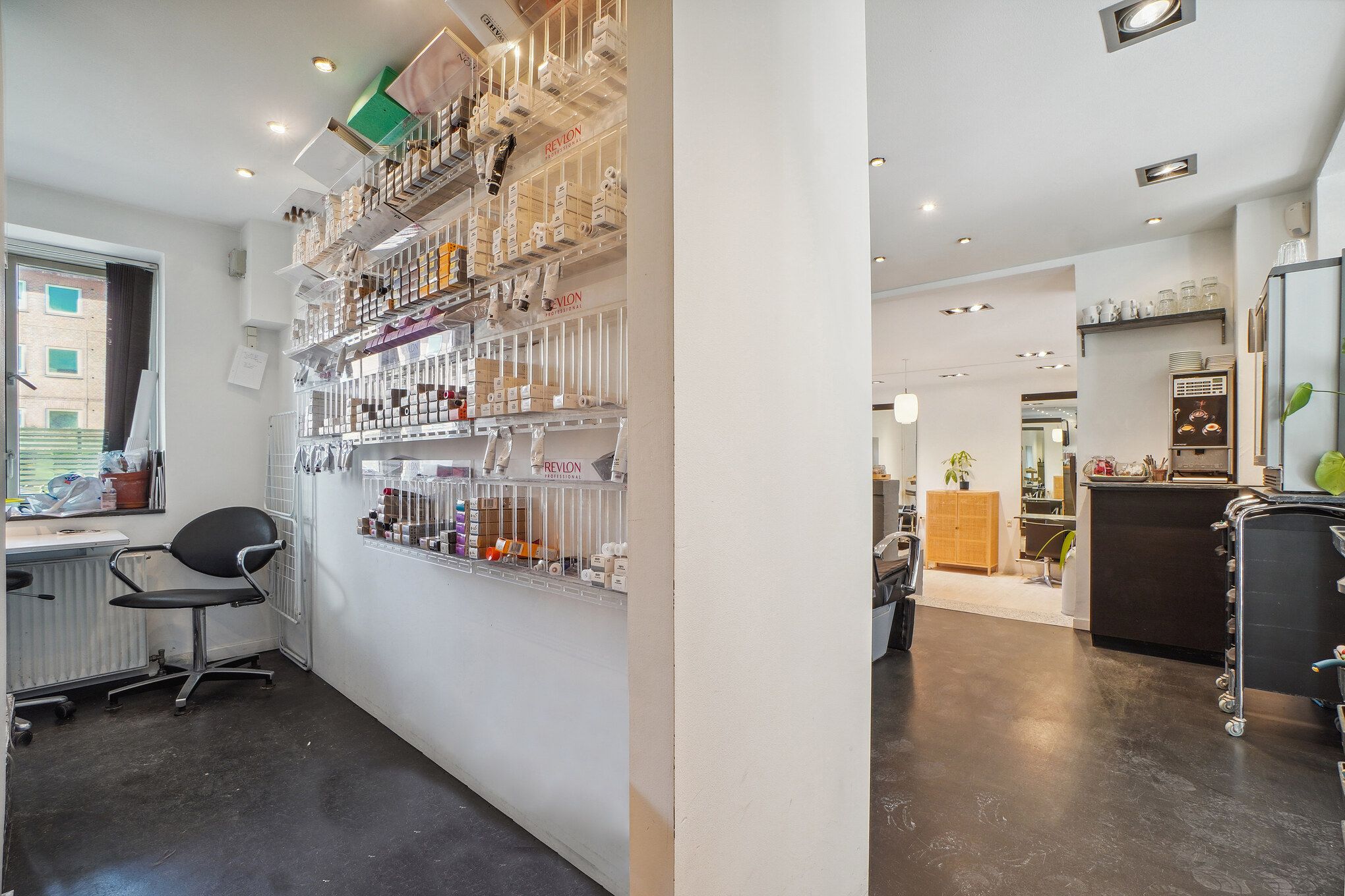 Frisørsalon med høj hylde med diverse produkter og udstyr, moderne interiør, lyse gulv og vægge samt et skrivebord med en