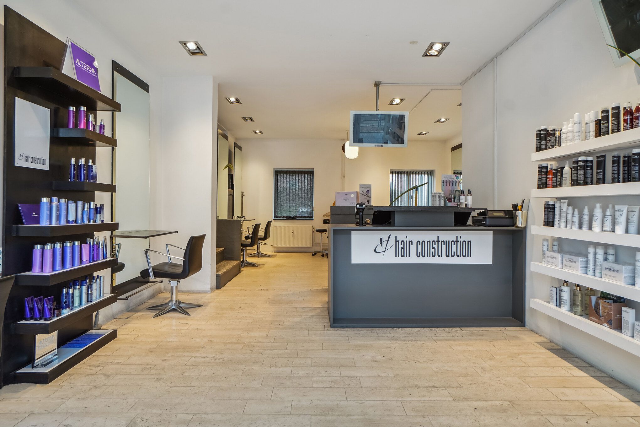 Indenfor en frisørsalon er der to barberstole og et salgsdisk med mærket Hair Construction. Der er to høje hylder med