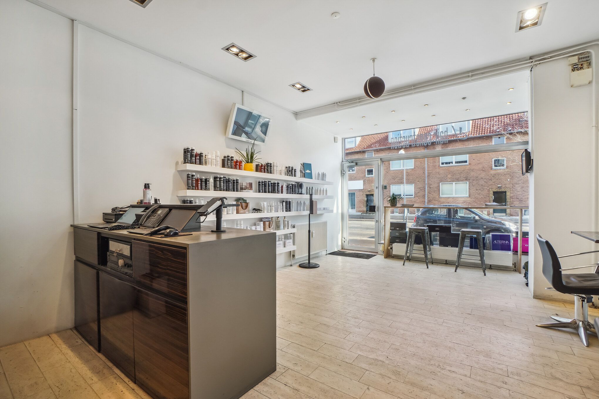 Frisørsalon med en lille salgsdisk, hvor der er plakater og produkter på hylderne og udendørs er der en bilparkeringsplads