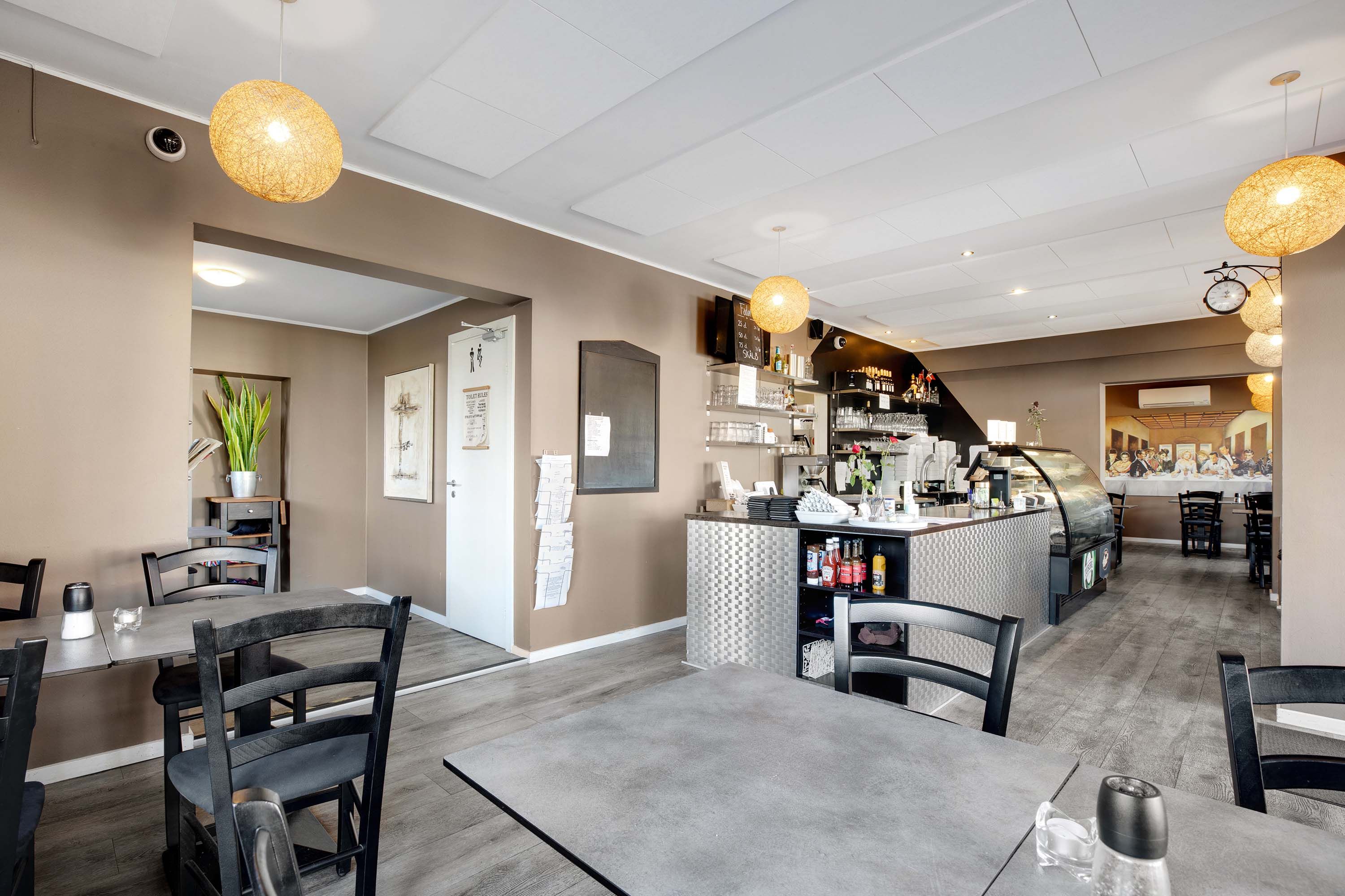 En restaurant med bord og stole, plante, lys, gulv, tavle og køkken.