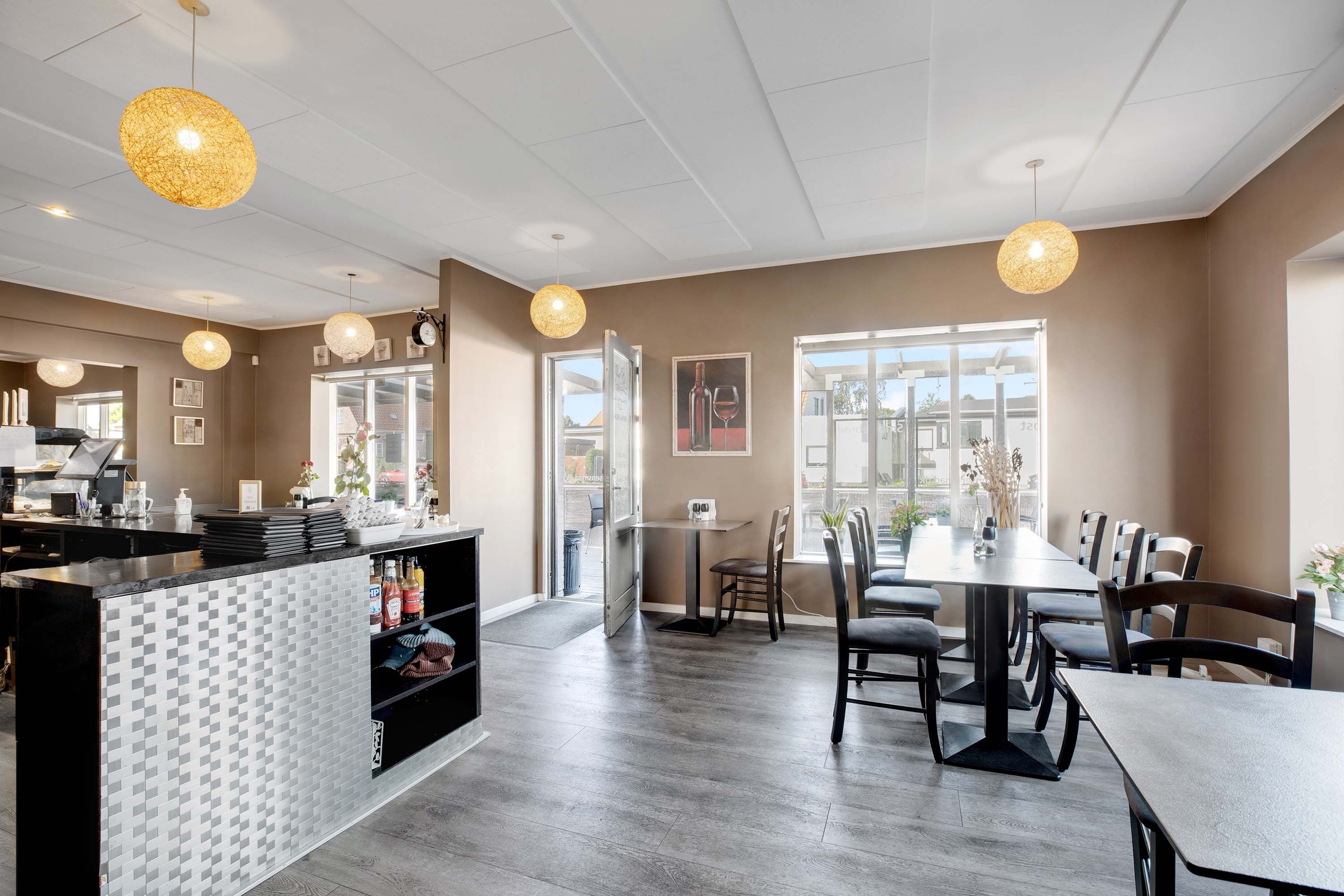 Restaurant med bord og stole, åben dør, lyskroner, sofaer, stole, servietter, drikkevarer og plante i vinduet