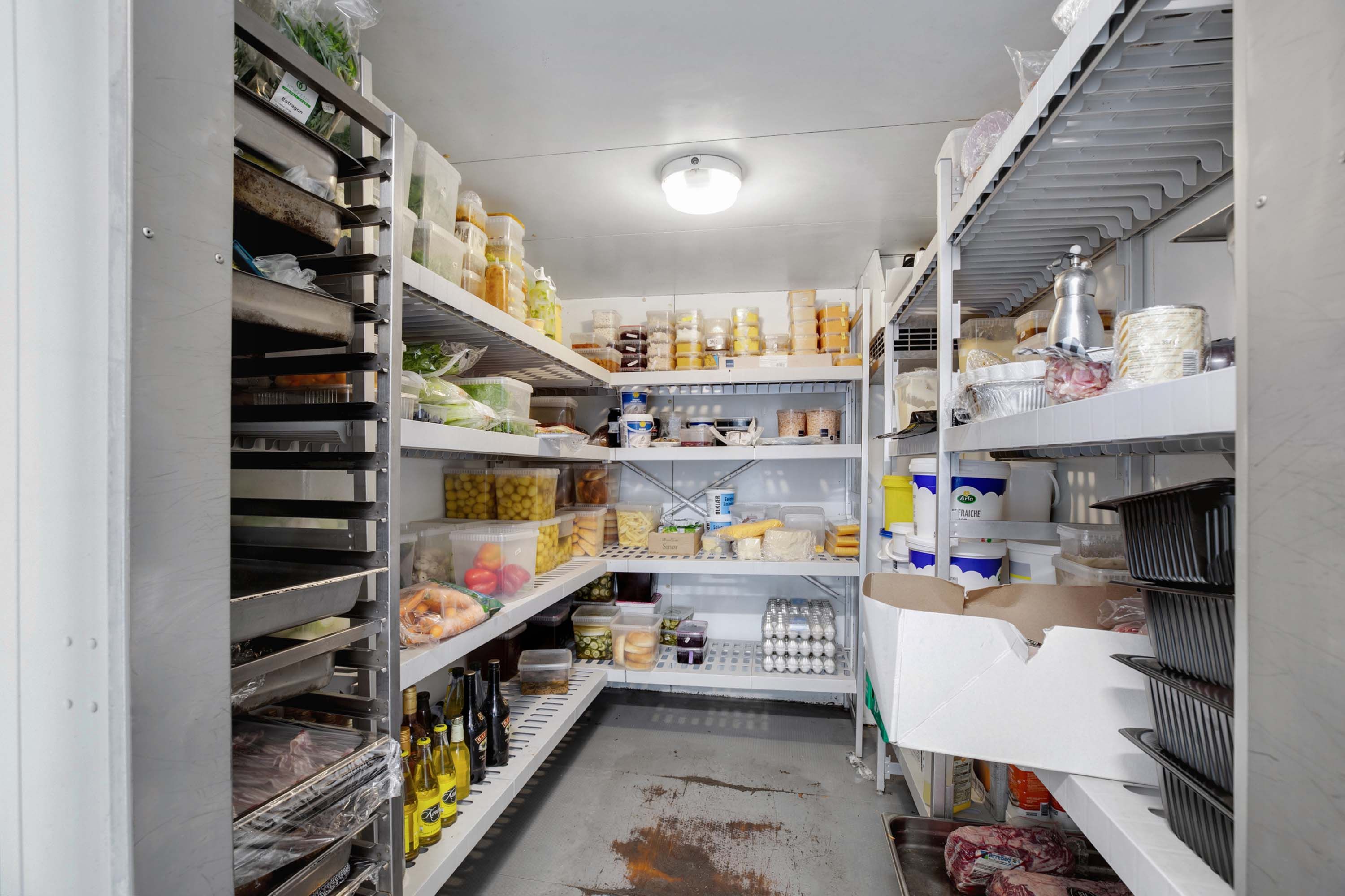 Kølekammer med flere hylder fyldt med mad og drikkevarer, bl.a. kød, grøntsager, æg, mælk, olivenolie og vin
