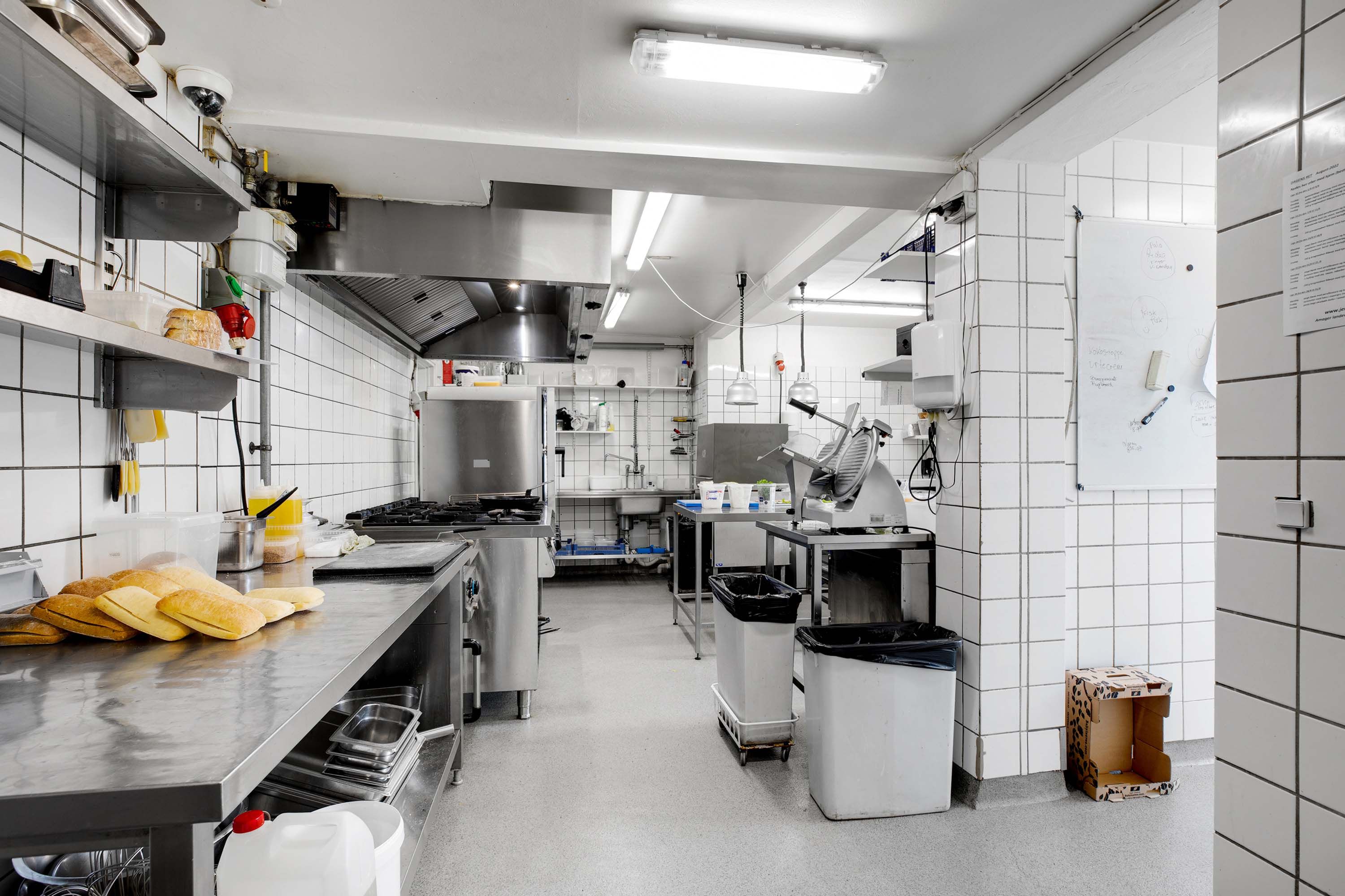 Et moderne køkken med flere apparater, skraldespande og hvidt flisede vægge. En stor ovn og et apparat til at sælge mad. En