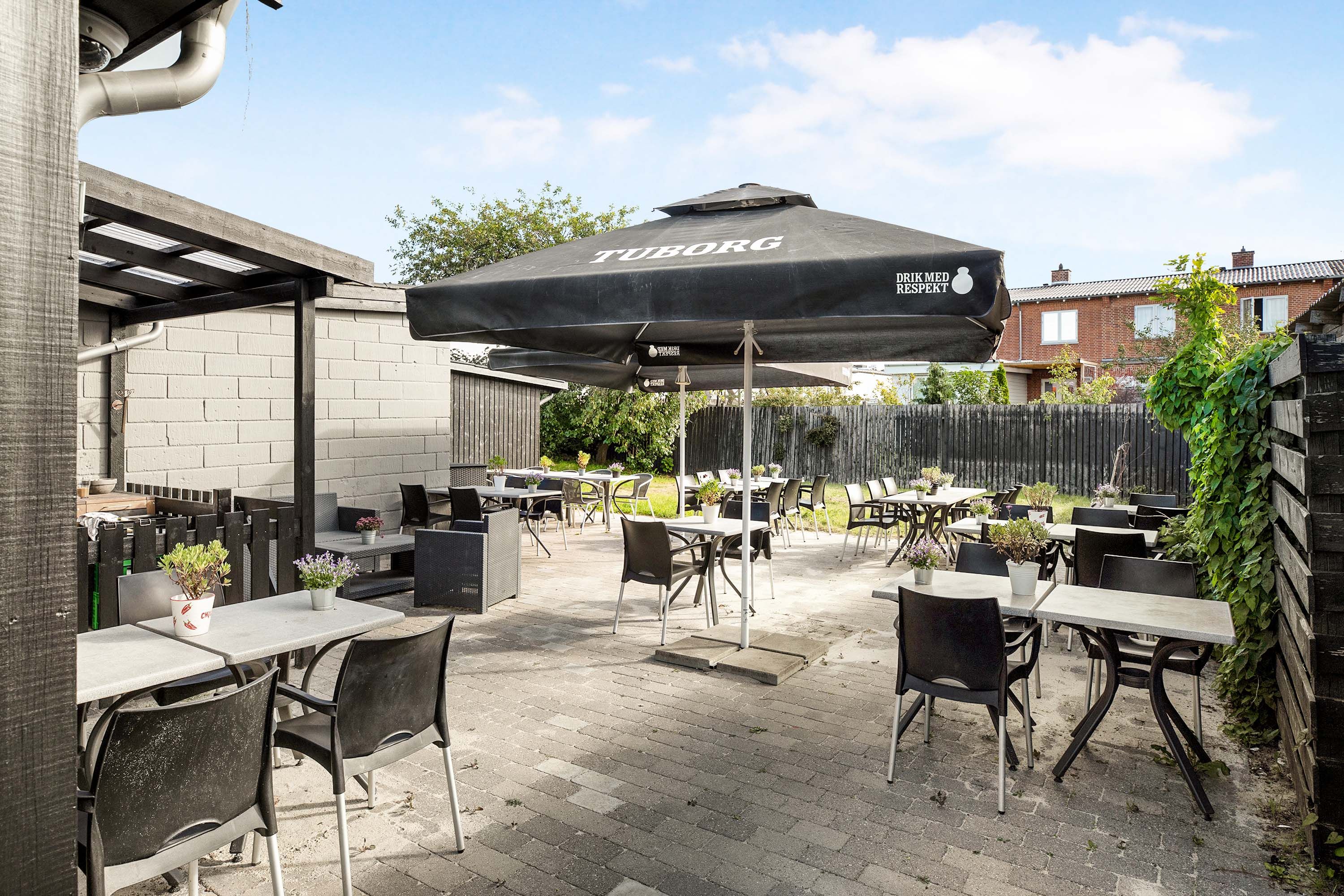En restaurant med flere borde og stole udenfor under et stort paraplymærke Tuborg og et træskur med planter