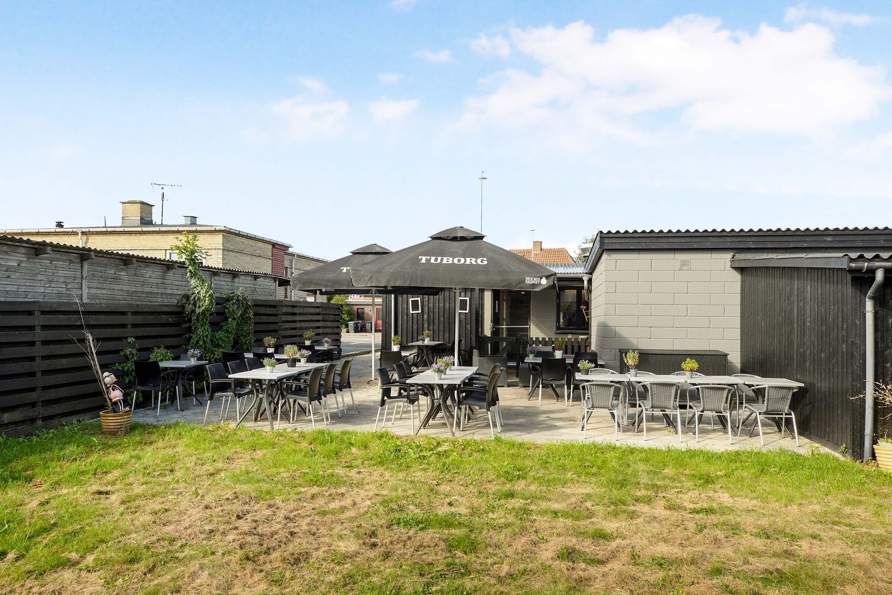 Udenforservering med flere borde og stole samt parasoller og blomster. Der er en terrasse med græs og buske.