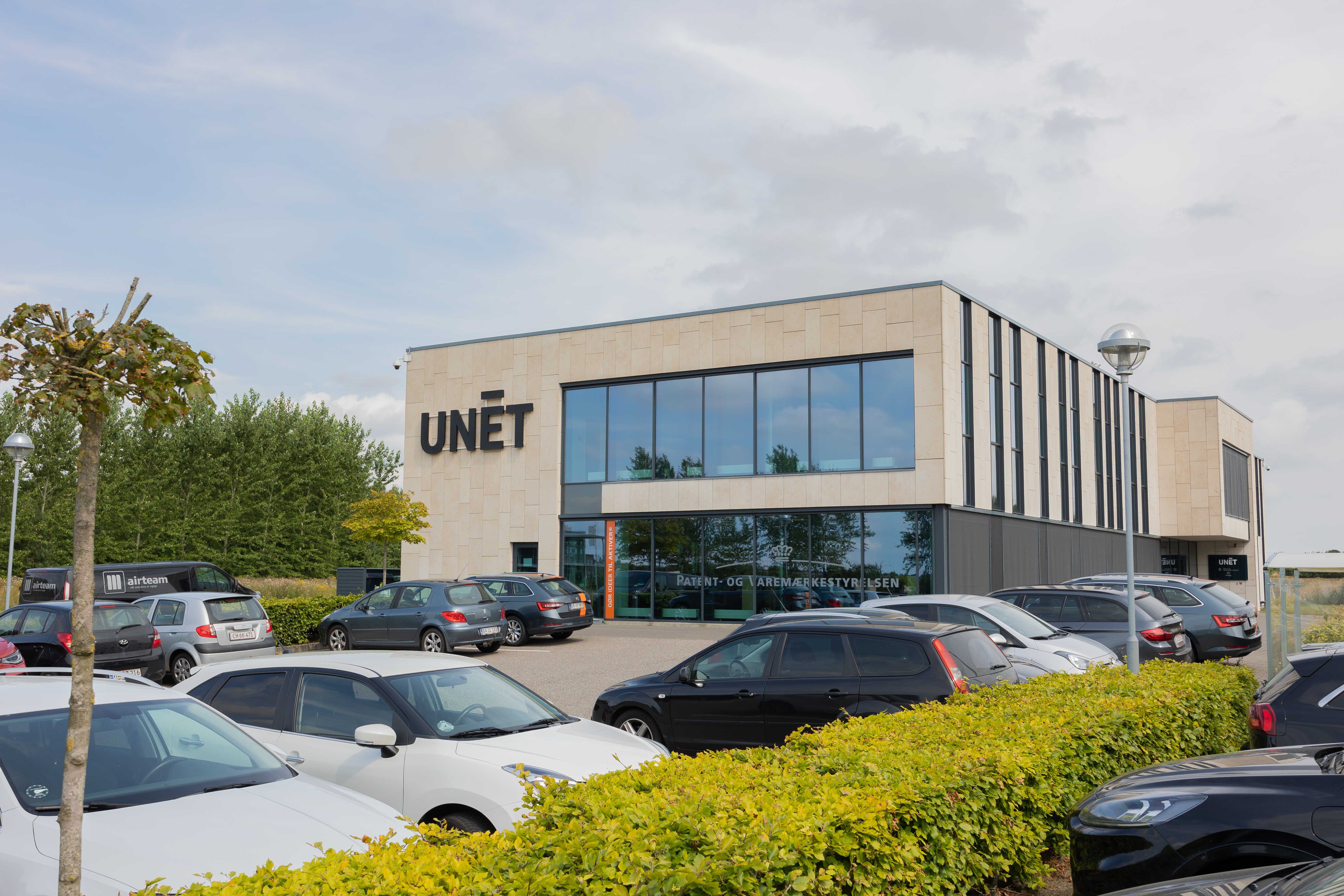 Moderne kontorbygning med logoet UNĒT. Foran bygningen er der en parkeringsplads med flere parkerede biler. Der er grønne