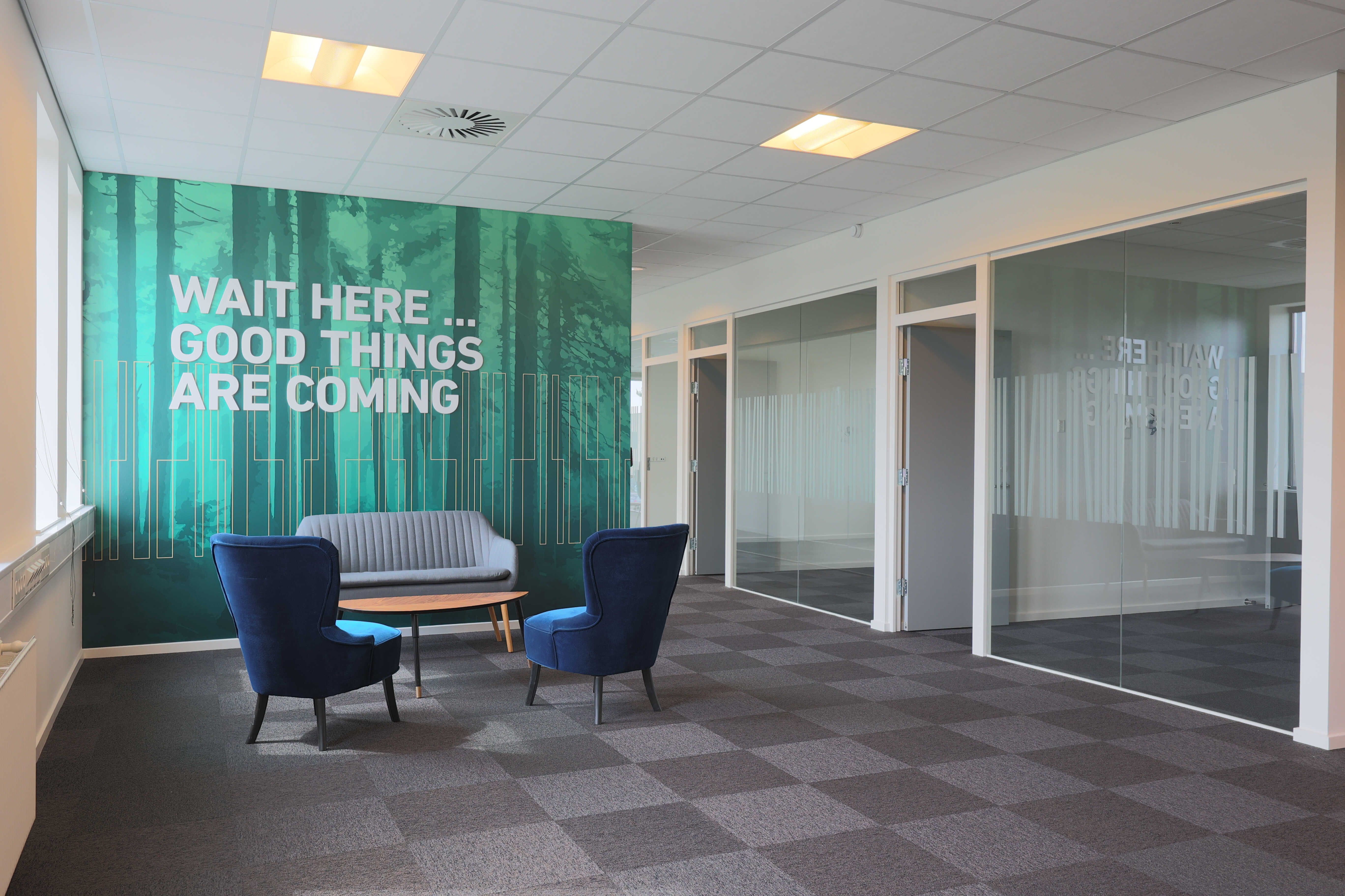 Et mødelokale med skrift på væggen, som lyder "Wait here... Good things are coming" samt to blå stole og et bord