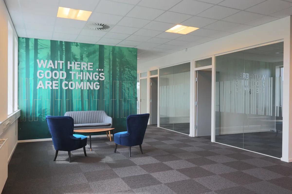 Et mødelokale med skrift på væggen, som lyder "Wait here... Good things are coming" samt to blå stole og et bord