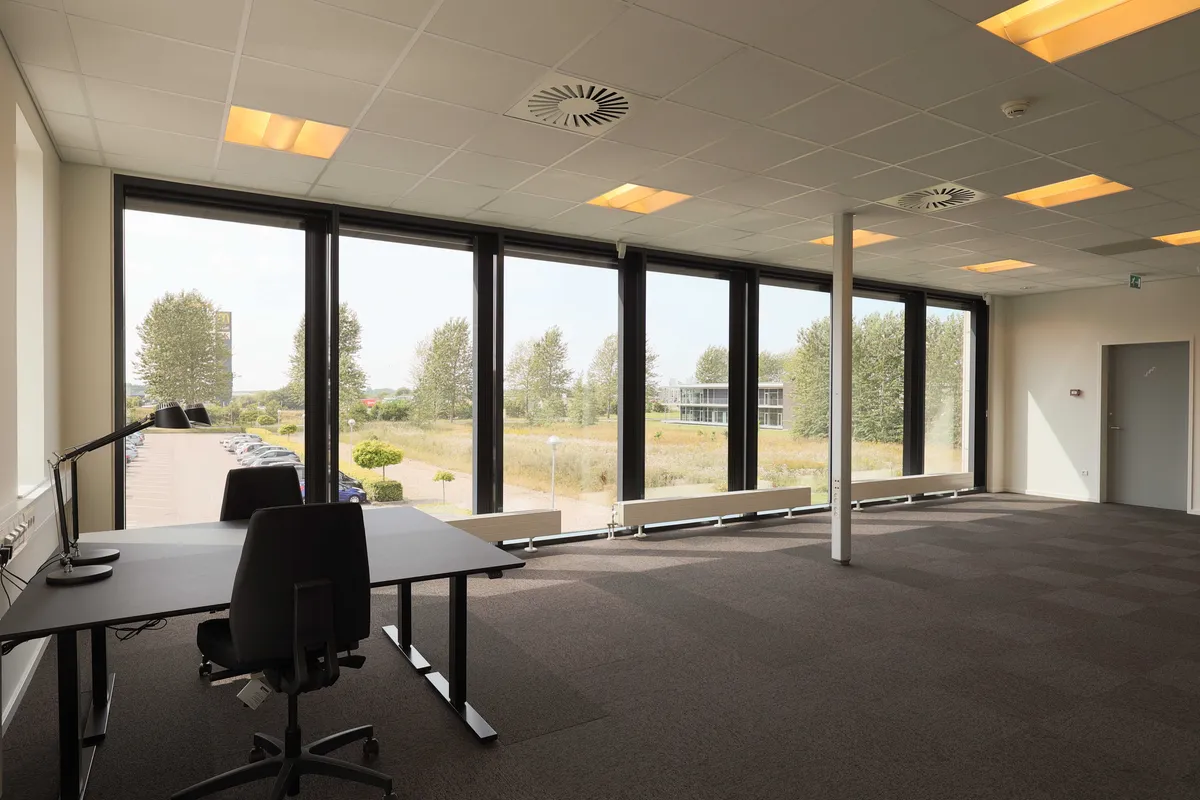 Lyst kontor med trægulve, skrivebord og stol samt store vinduer med udsigt til parkeringsplads og grønne områder