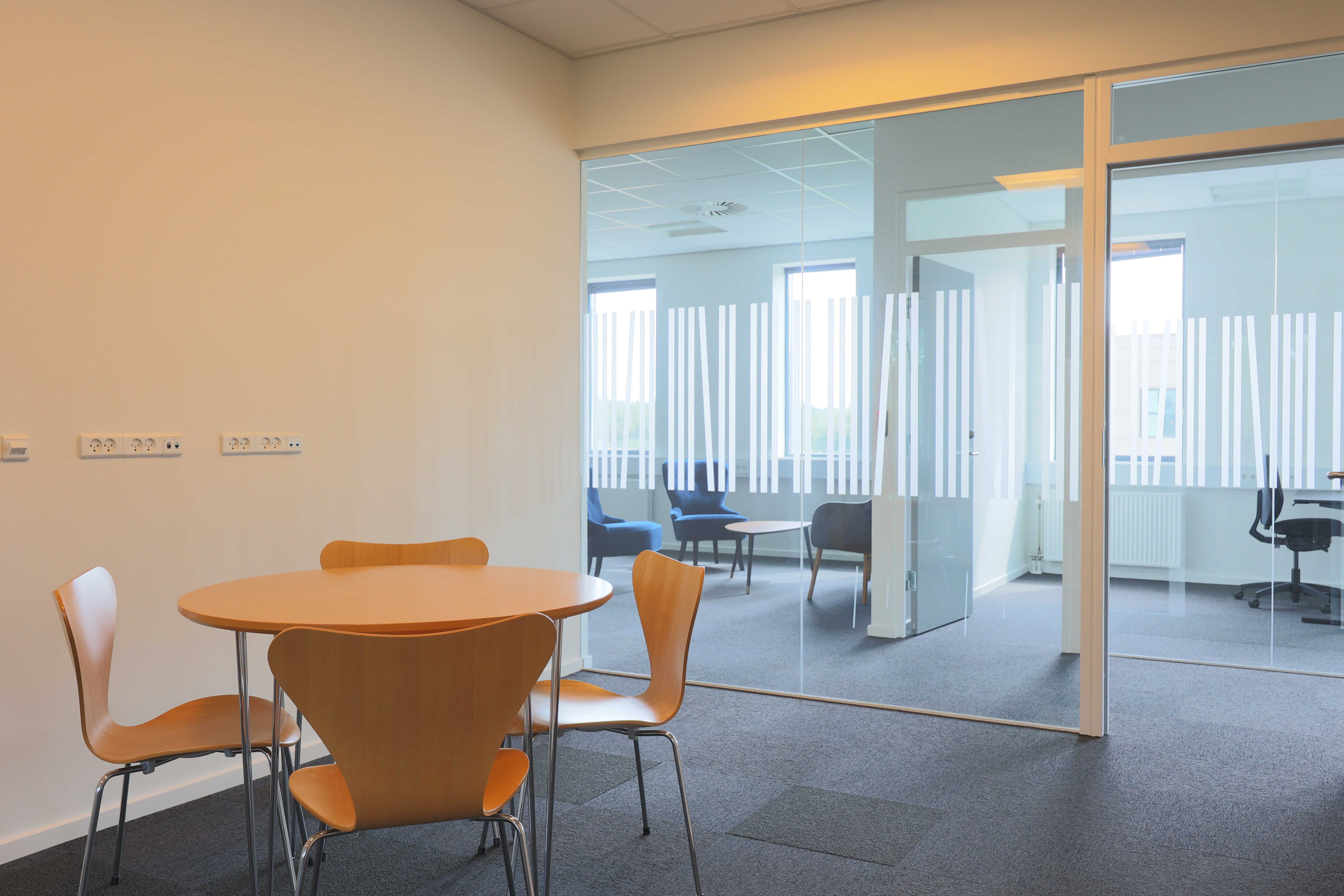 Kontorrum med rundt bord og fire stole samt et glasadskillelsesvælv, der giver udsigt til et lysere rum med blå møbler og