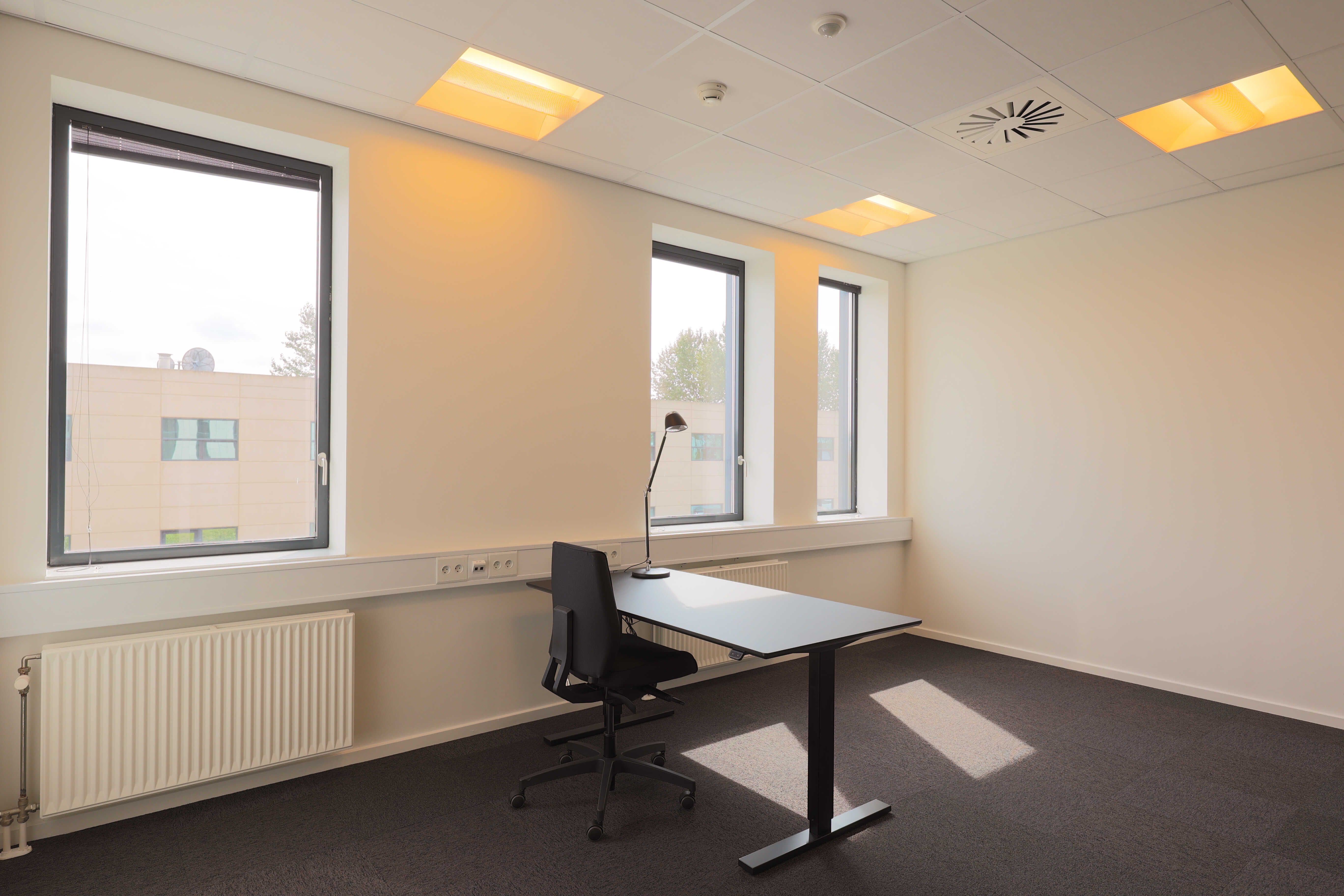 En tom kontorkontor med en høj hvid væg og en skrivebordsstol med lampe placeret i spidsen af skrivebordet. Et radiator er