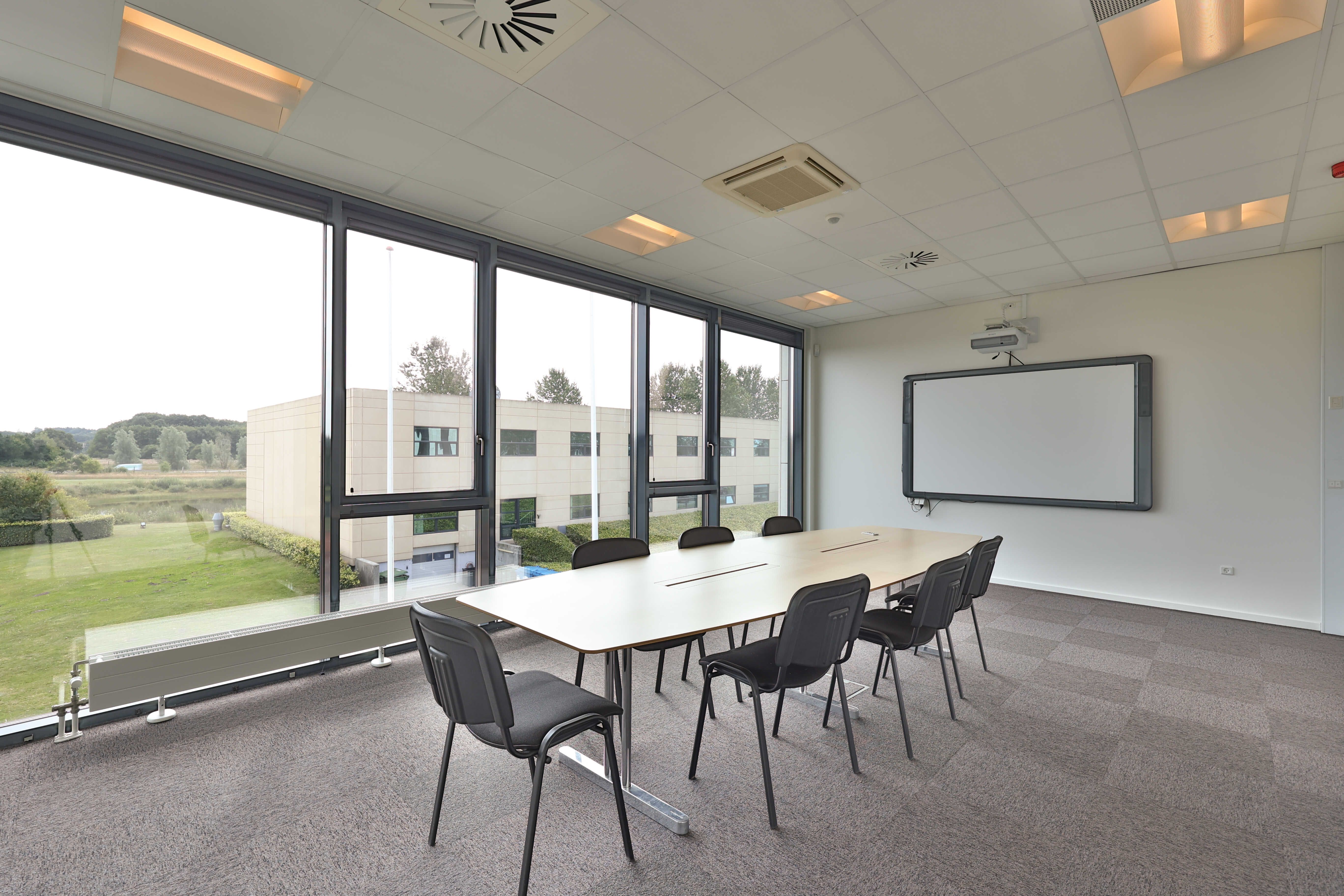 Lyse møderum med store vinduer og lysegule vægge, hvor der er et stort hvidt projektionsskærm og moderne møbler med glasbord