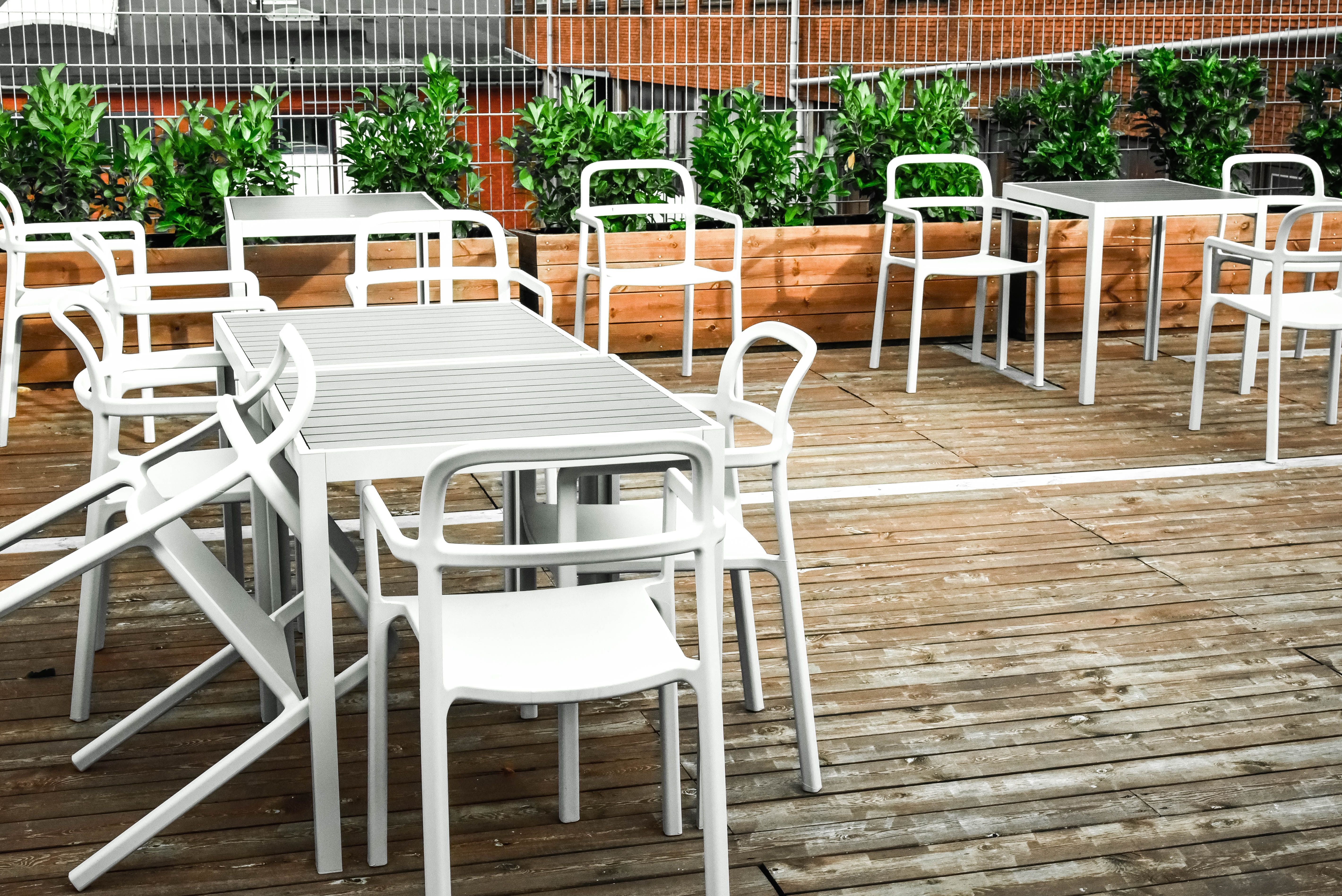 Forskudte hvide stole og borde på en terrasse med udsigt til planter og bygninger i baggrunden