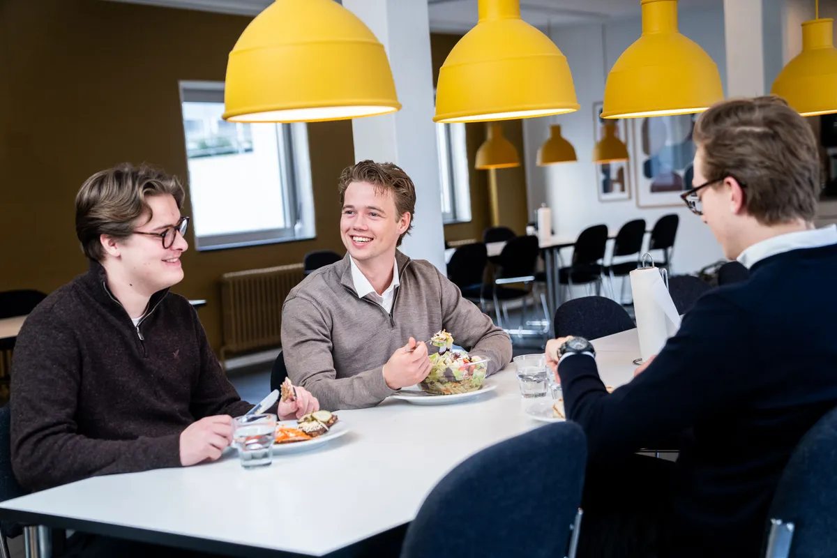 Tre unge mænd sidder og snakker og spiser i en stor, veludstyret kontorrestaurant med lysegult gulv og gule lysekroner