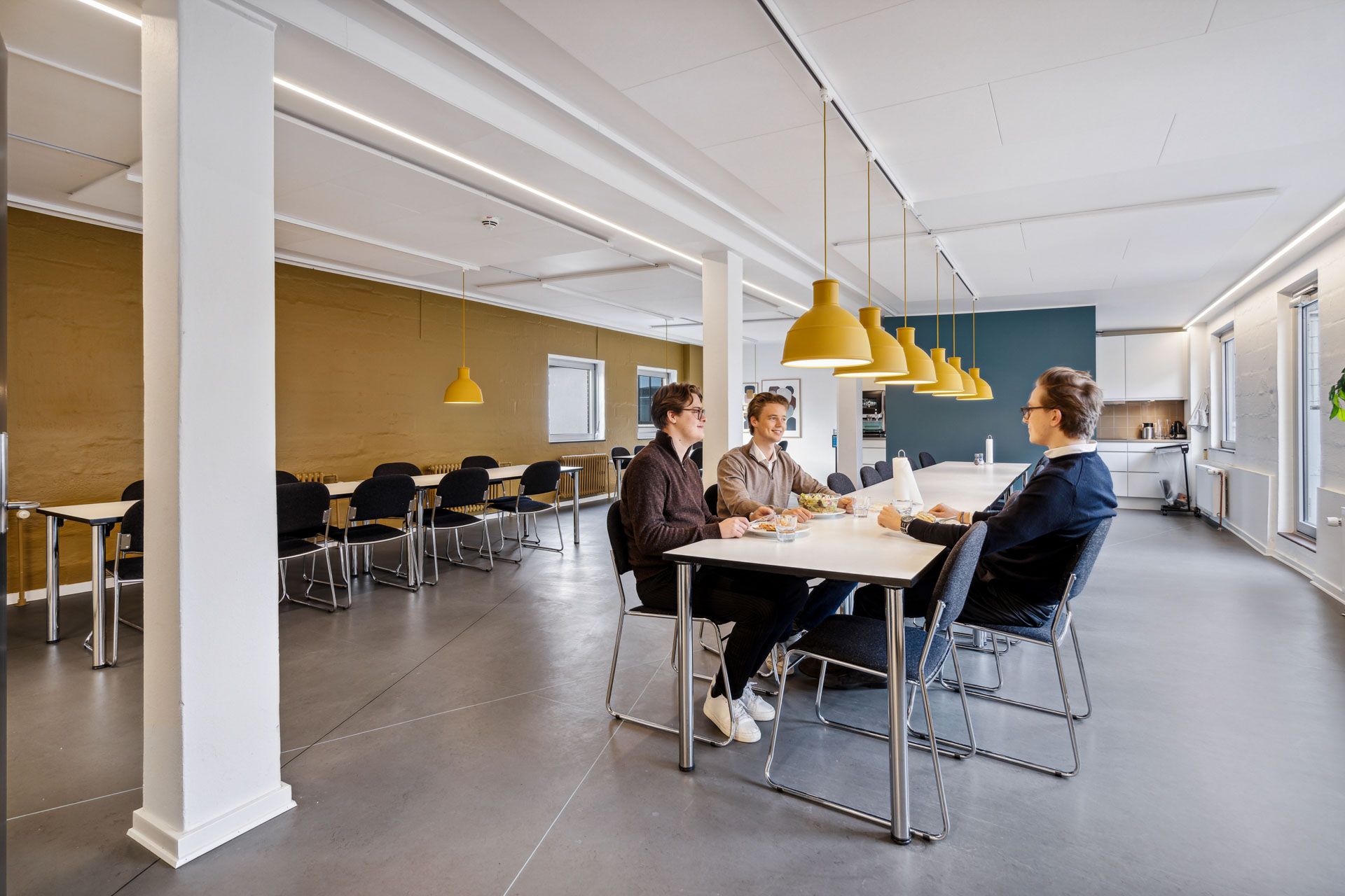 En stor fælles spisesal med bord og stol og tre unge mænd, der sidder og snakker med hinanden