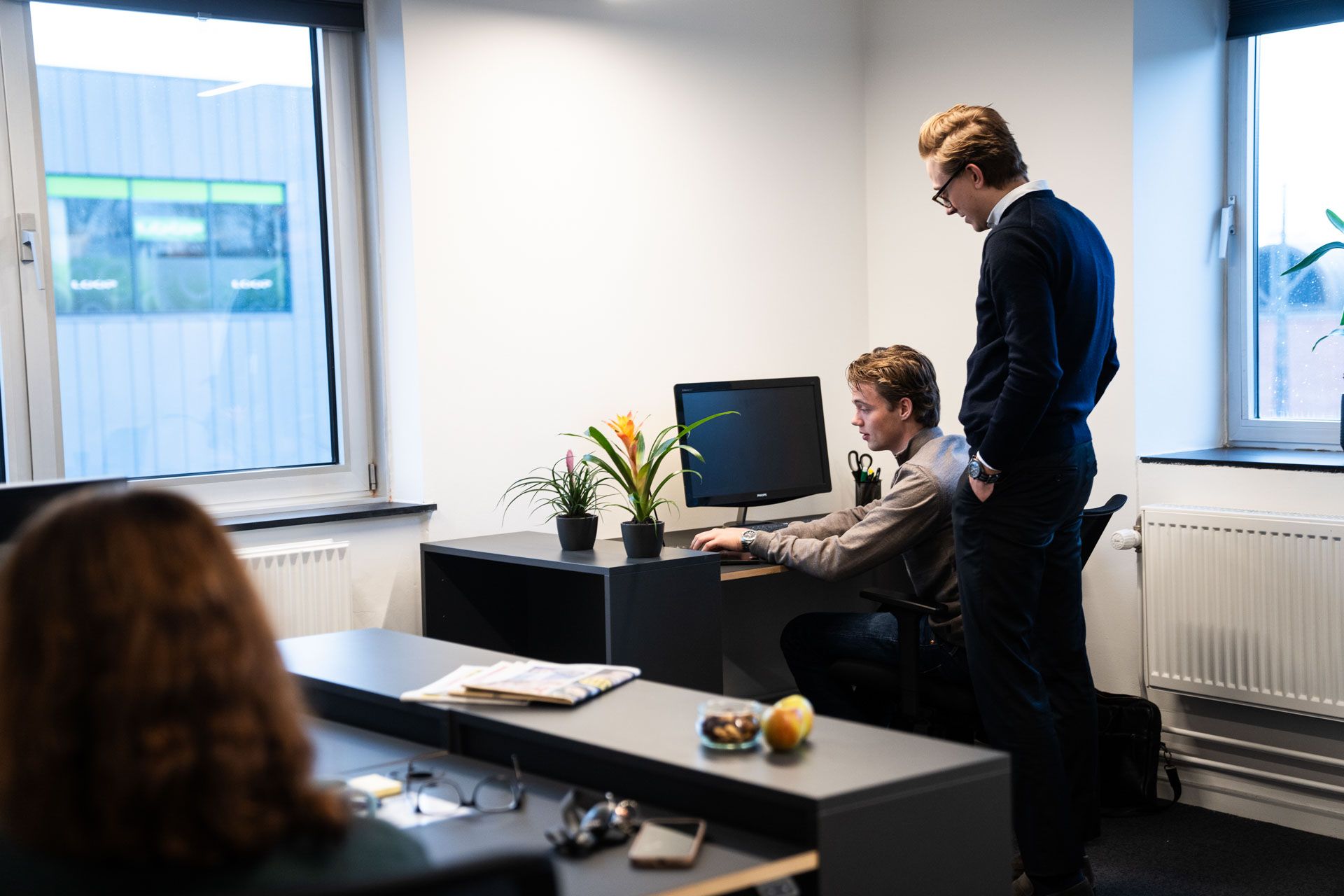 To mænd står og snakker i et kontor med vinduer og plante på skrivebordet