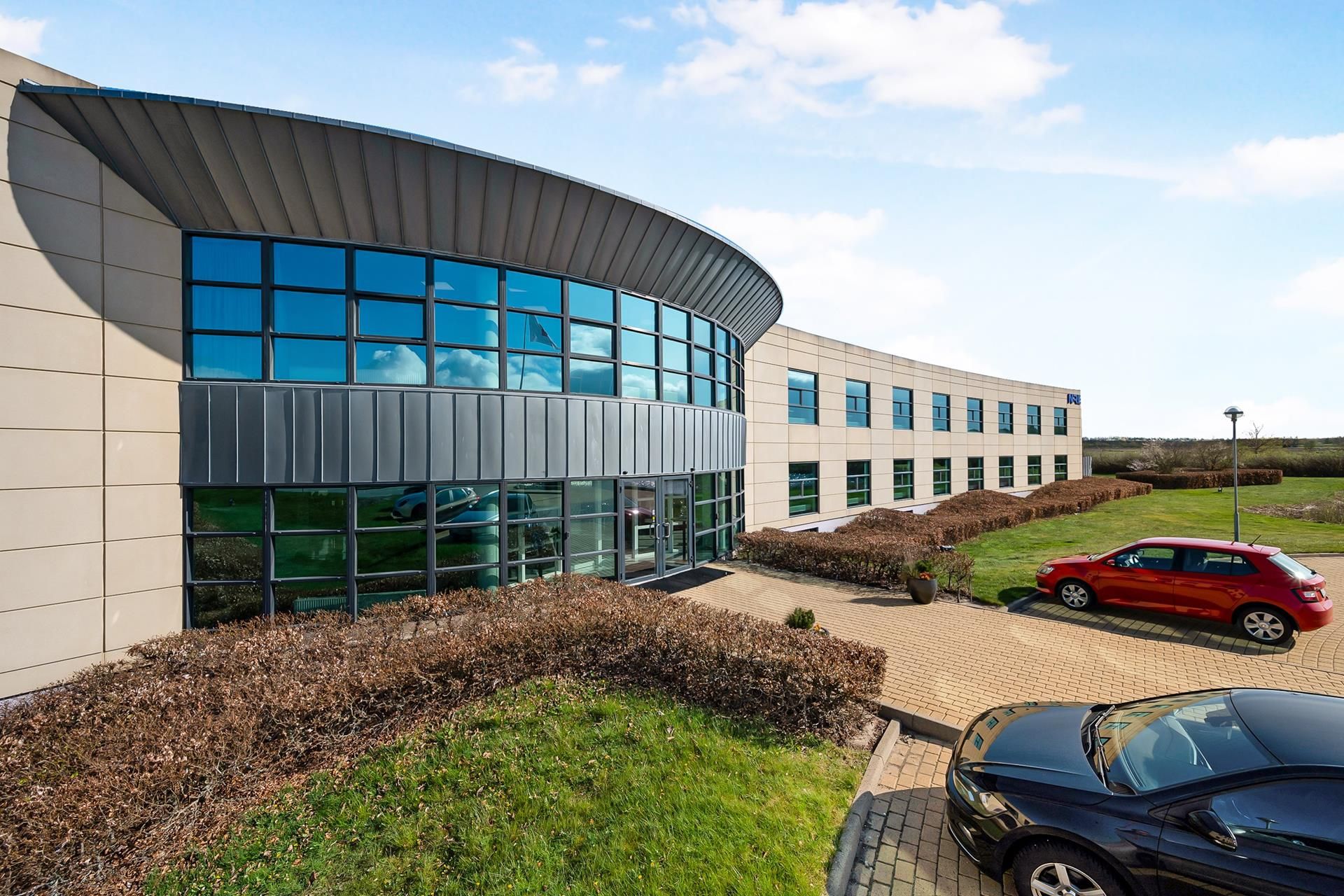 Kontorbyggeri med en glas facade og to biler parkeret udenfor. Lys og skyer i baggrunden og et grønt græsplæne.