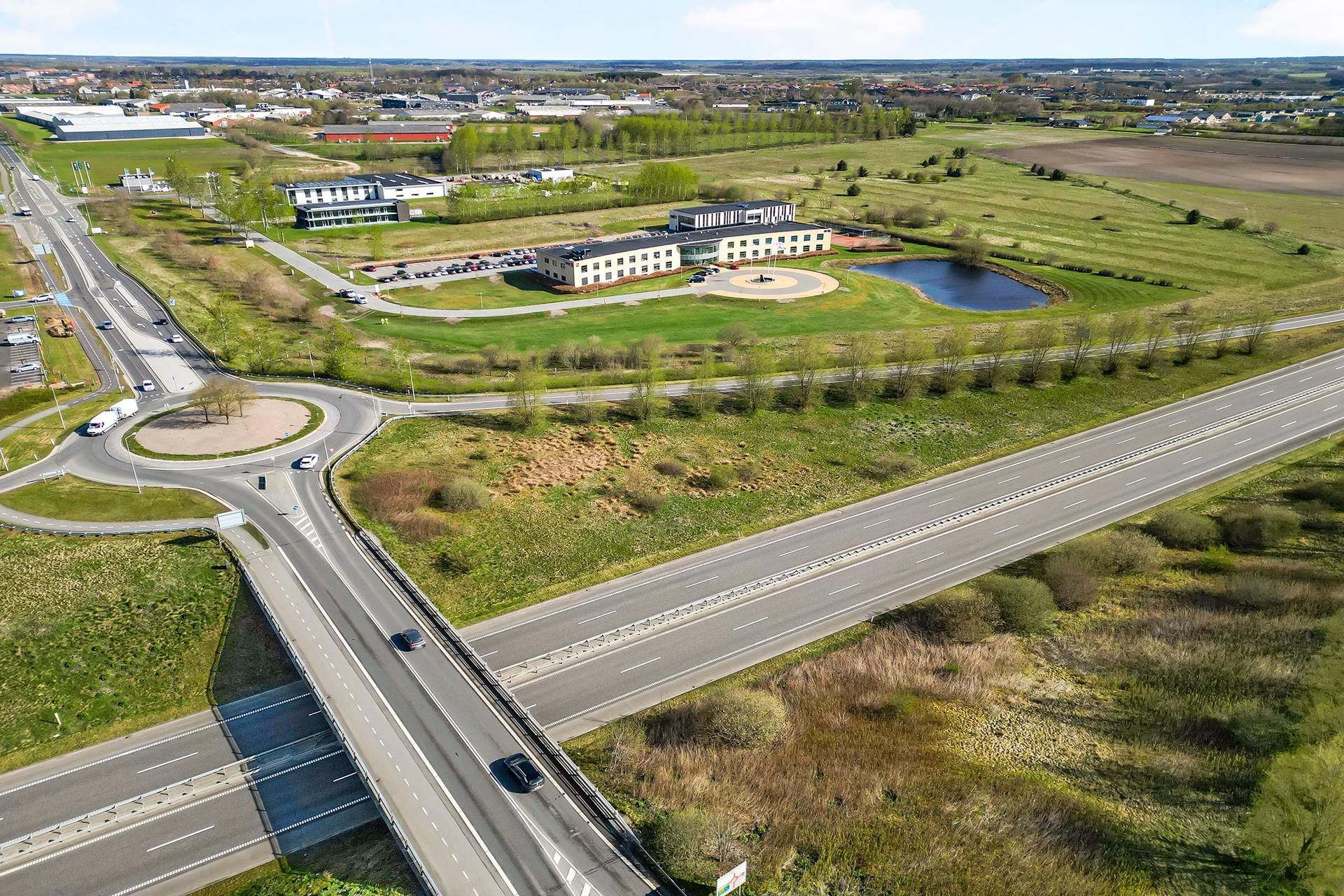 Vejkryds med svinge og motorvej med biler, grønne marker og sø samt bygninger i baggrunden