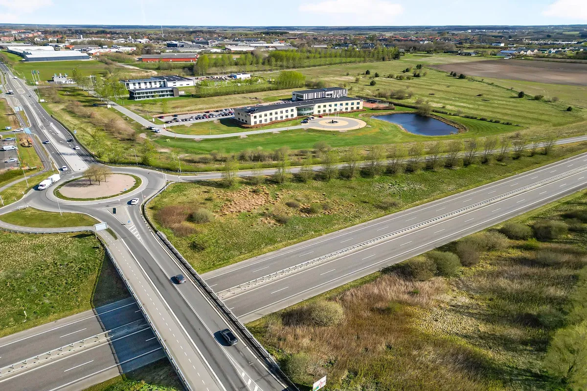 Vejkryds med svinge og motorvej med biler, grønne marker og sø samt bygninger i baggrunden