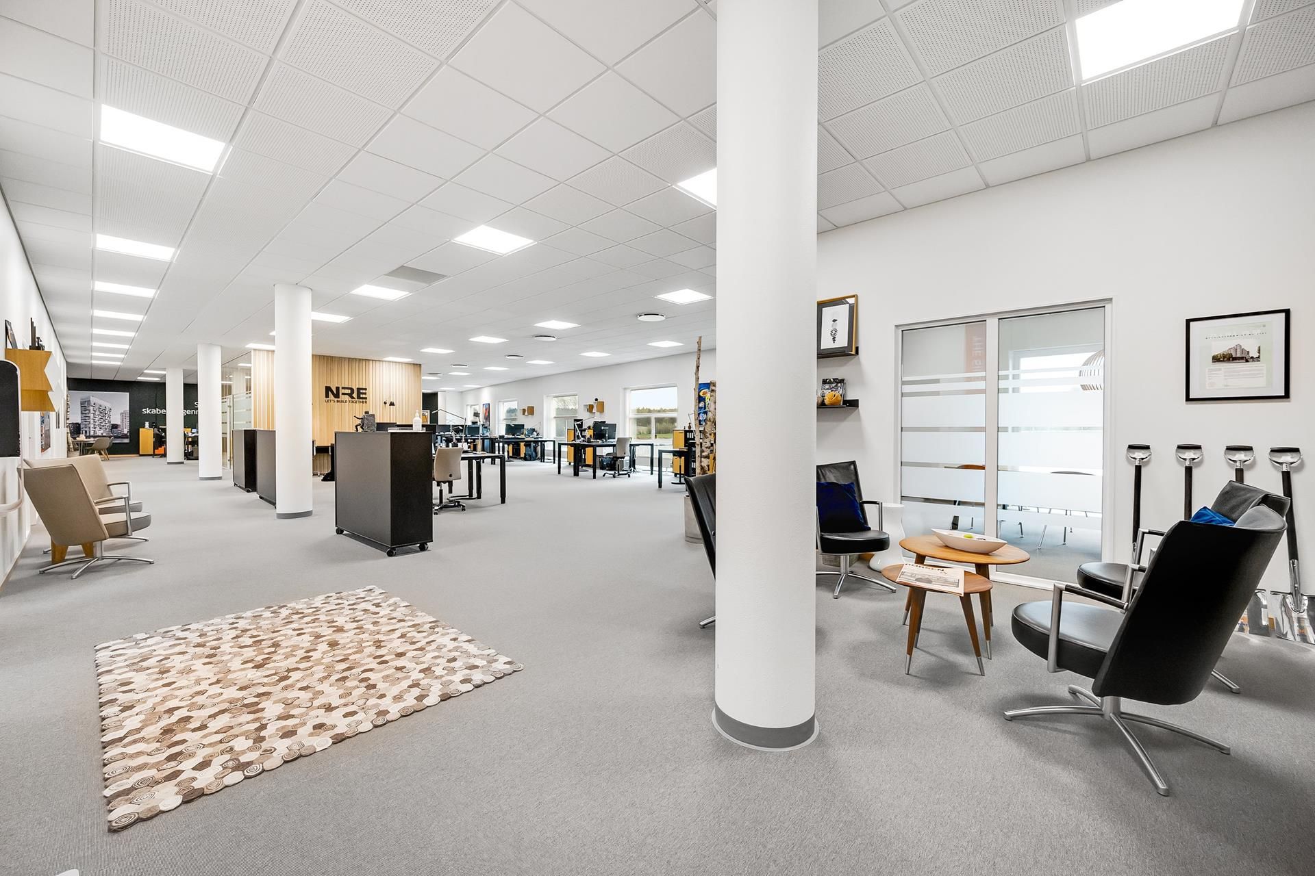 Kontorlokaler med møbler og indretning, hvor skriveborde og stole er placeret langs væggen, og der er en stol med et bord og