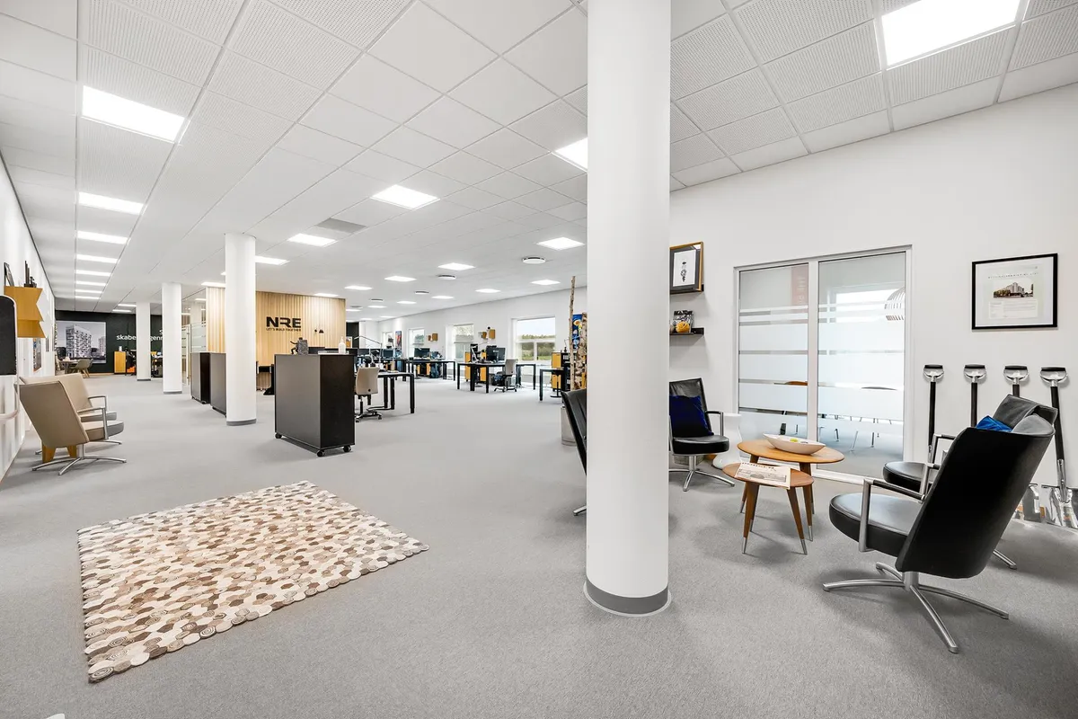 Kontorlokaler med møbler og indretning, hvor skriveborde og stole er placeret langs væggen, og der er en stol med et bord og