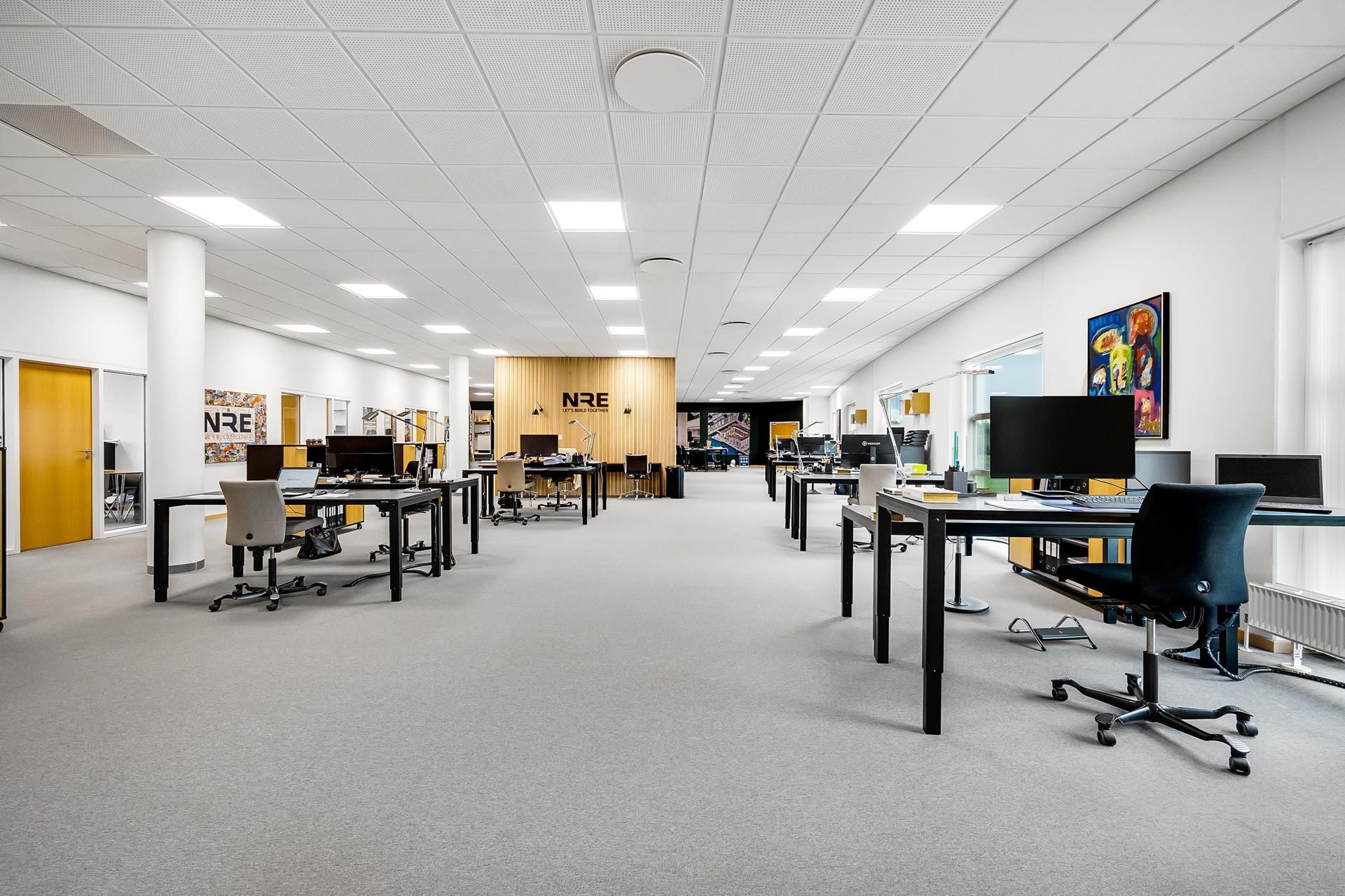 Kontor med lyse møbler og gulvbelægning og store vinduer med udsigt til udendørsområder.