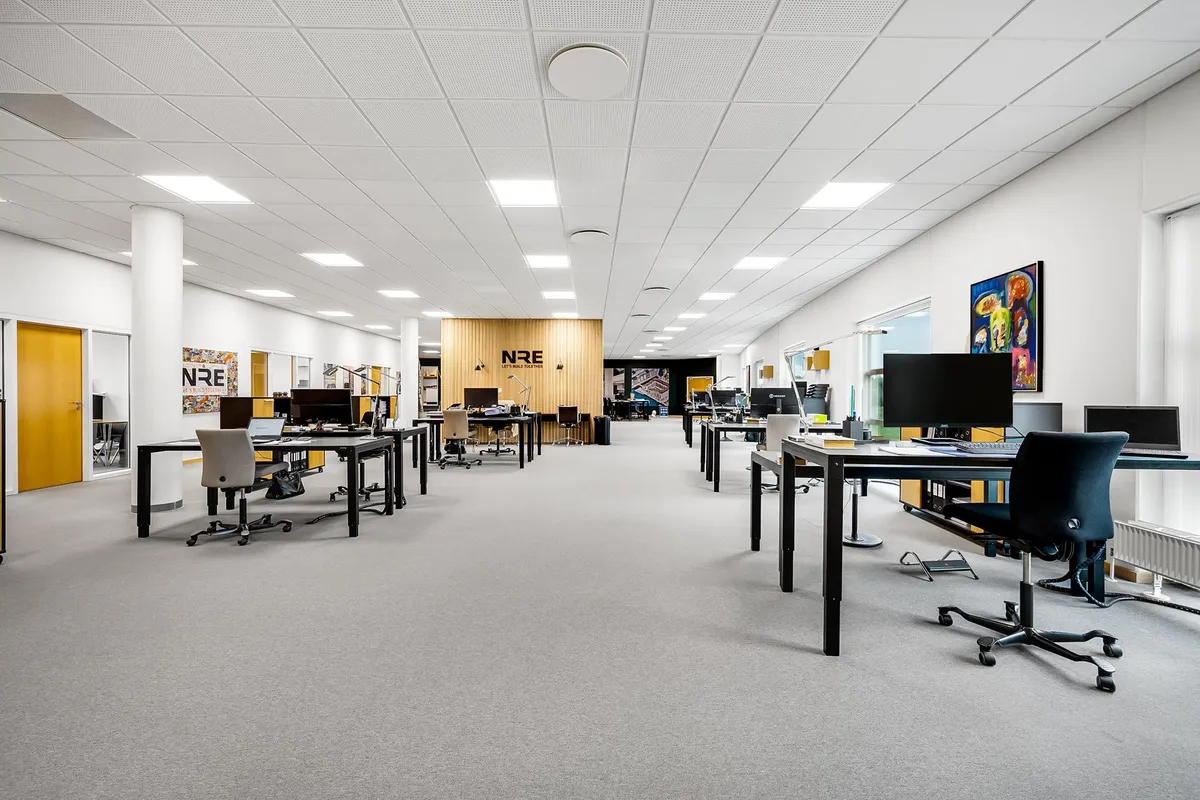 Kontor med lyse møbler og gulvbelægning og store vinduer med udsigt til udendørsområder.