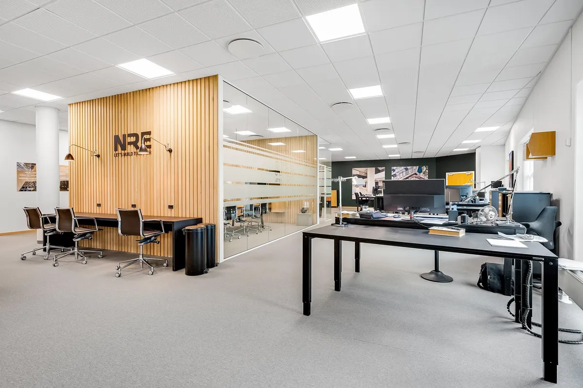 Kontor med moderne design, lyse lofter, trævægge, og møbler med høj kvalitet og simpel stil. Væggen har en stor logo og