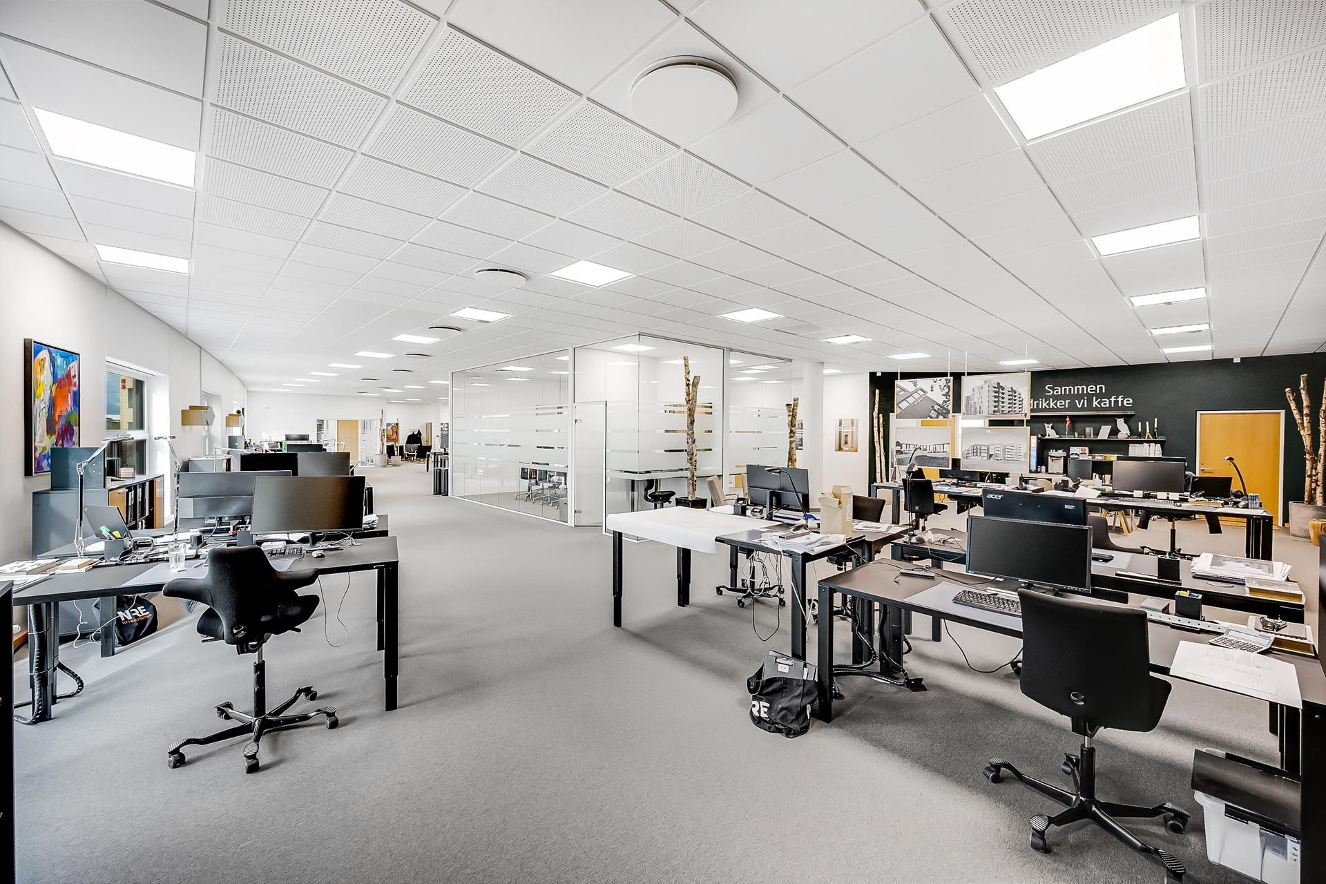 Kontor med computere, stole, borde og tavle med tekst og billeder. Der er lyse lofter med indbygget belysning og hvide vægge.