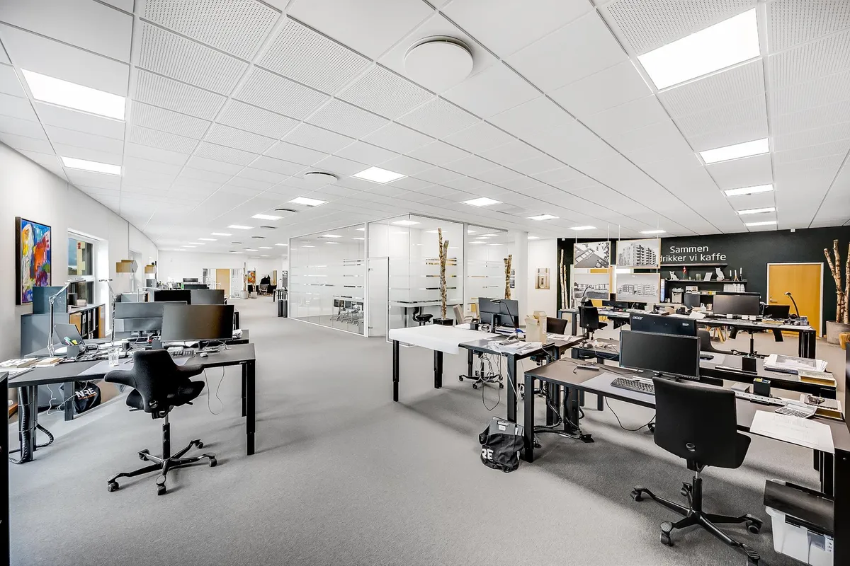 Kontor med computere, stole, borde og tavle med tekst og billeder. Der er lyse lofter med indbygget belysning og hvide vægge.