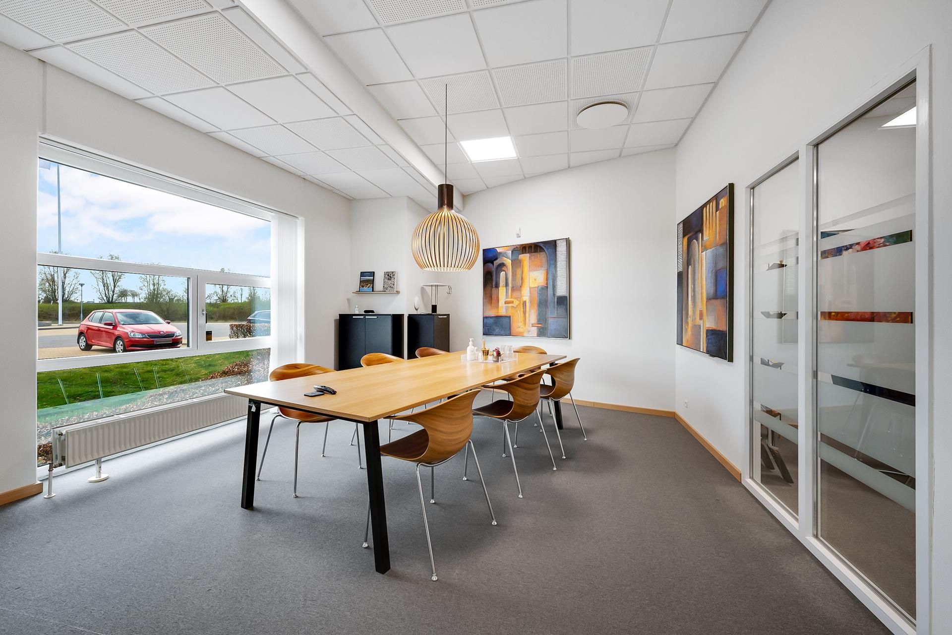 Kontorrum med store ruder og lyse lofter, møbler og vægdekorationer, udendørs parkeringsplads med biler og grønne træer