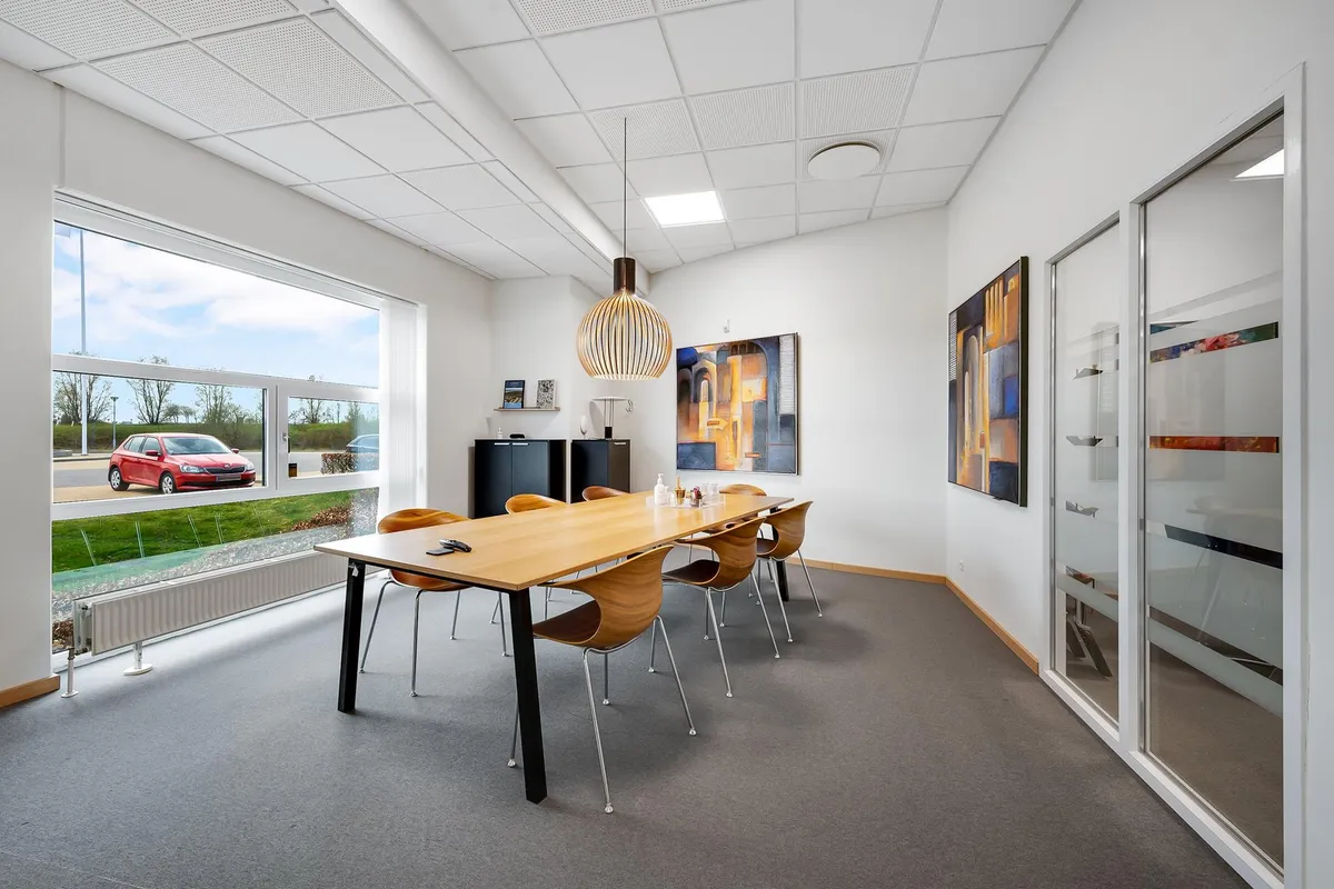 Kontorrum med store ruder og lyse lofter, møbler og vægdekorationer, udendørs parkeringsplads med biler og grønne træer