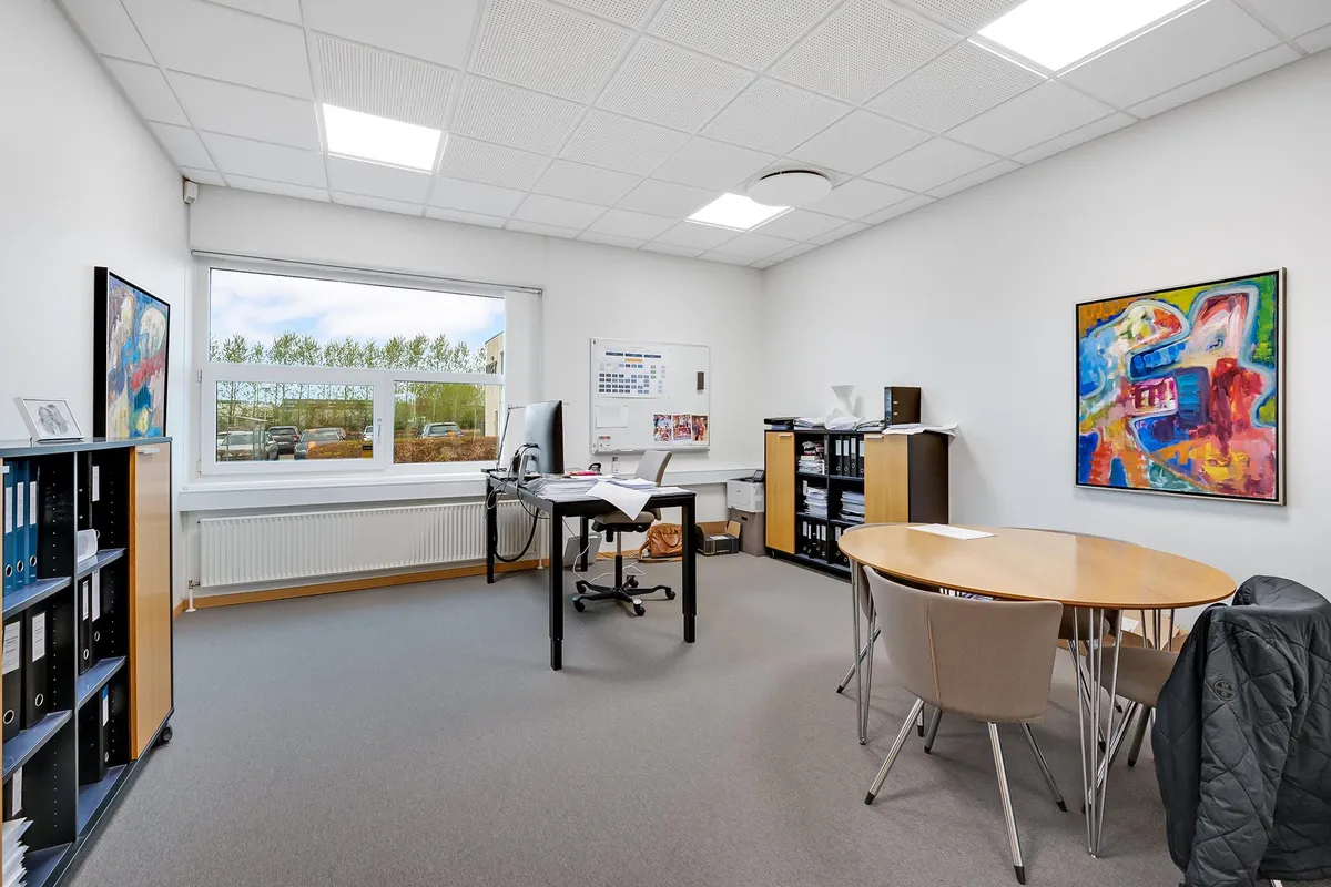 Kontor med runde borde, stole og skriveborde. Hvid væg og højt loft. Maleri og tv på væggen. Billeder og skriftligt