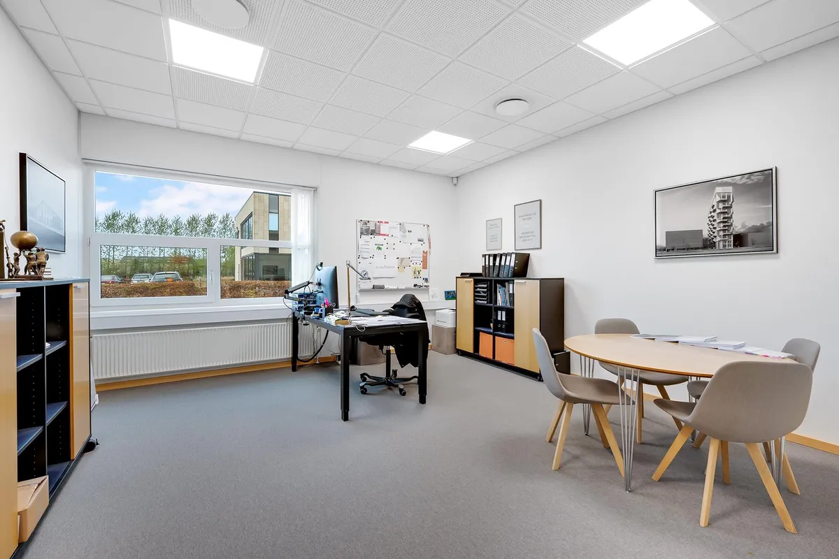 Kontor med lyse loftsbelægninger, højvinduer og moderne møbler. Bord med computer, stol og bordlampe. Væg med tavle og