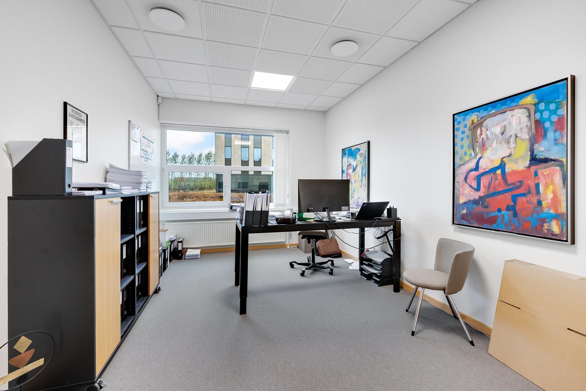 Et moderne kontor med en skrivebord, stol, computer, værktøj, kunstværk, højtalere, skriftbord og et lagerskab med papirer