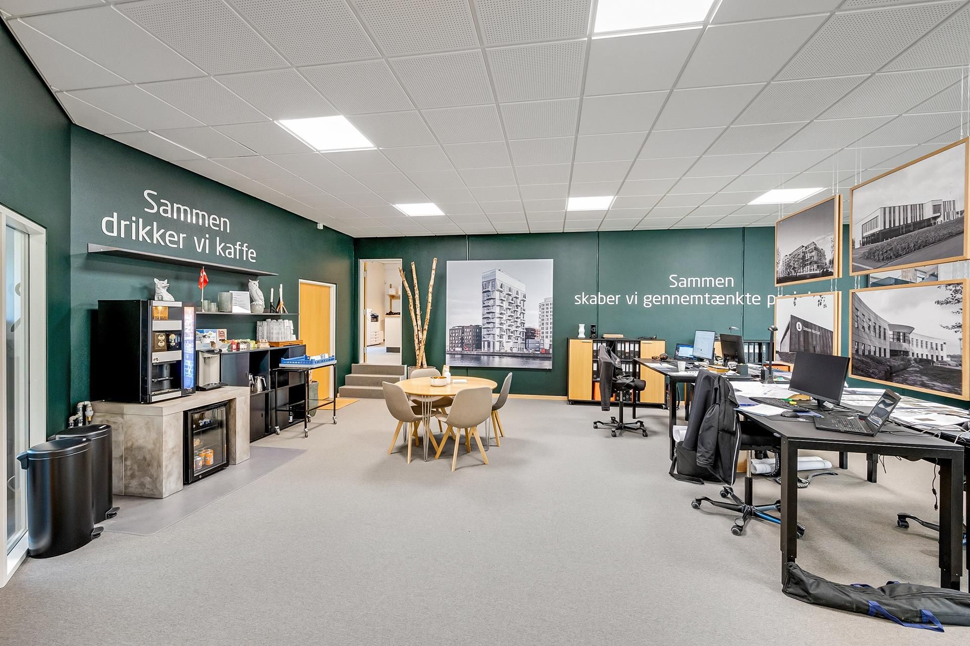 Kontorområde med lyse grønne vægge, et runde bord med stole og en kaffemaskine på væggen. Der er computere og stole placeret