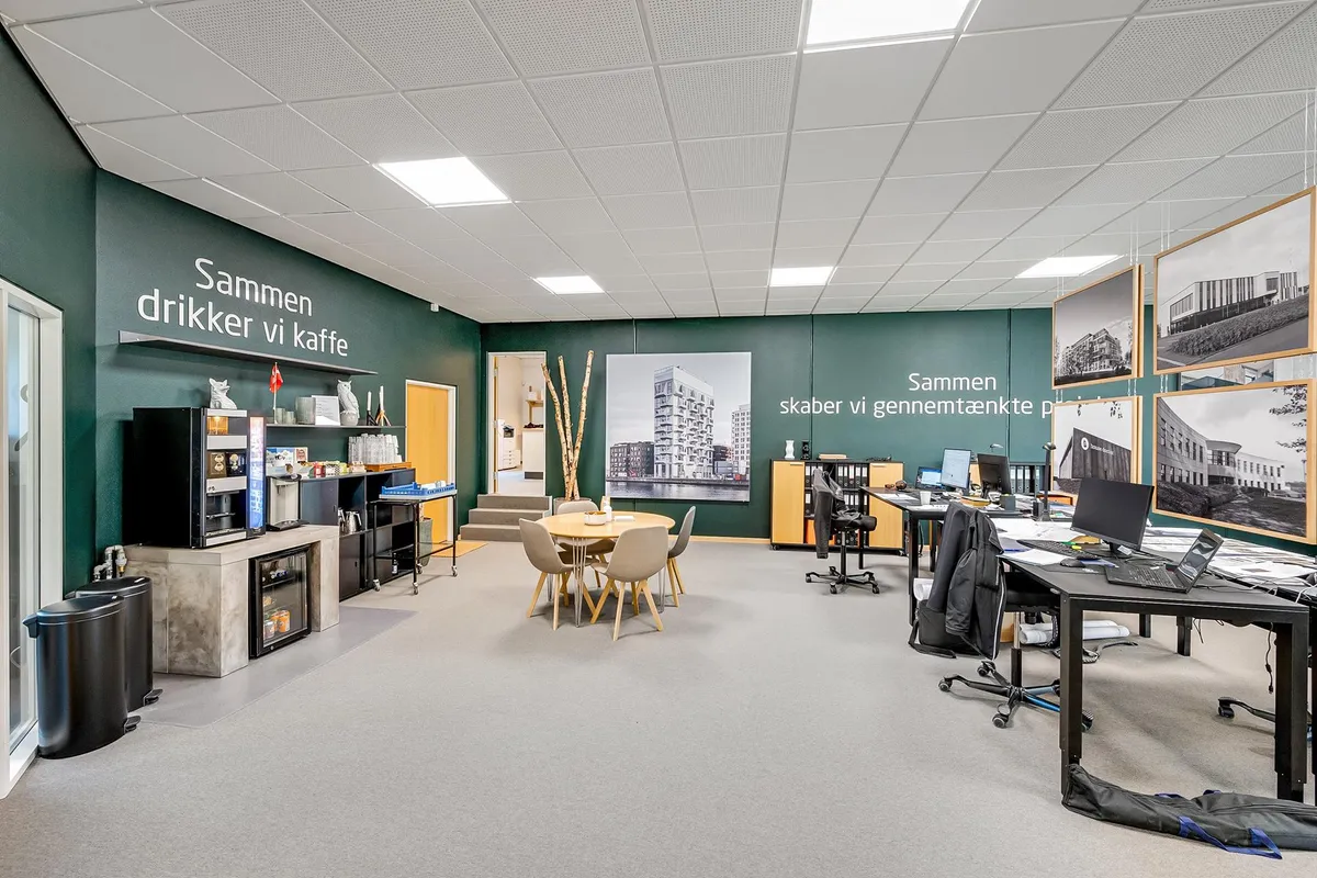 Kontorområde med lyse grønne vægge, et runde bord med stole og en kaffemaskine på væggen. Der er computere og stole placeret