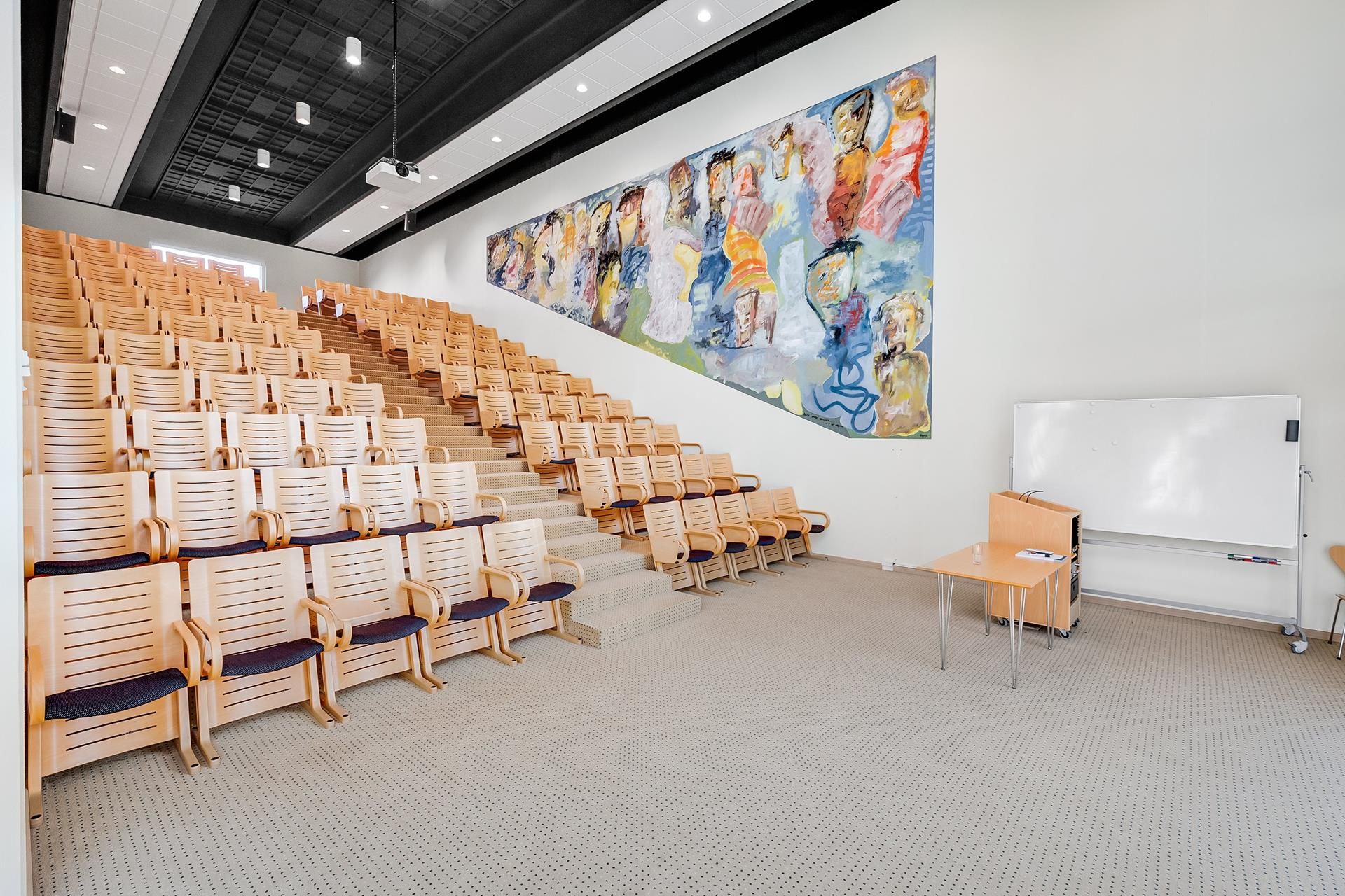Auditorie med træstole og puder og et stort maleri på væggen og hvide tavle med en lille bord og en stol i hjørnet