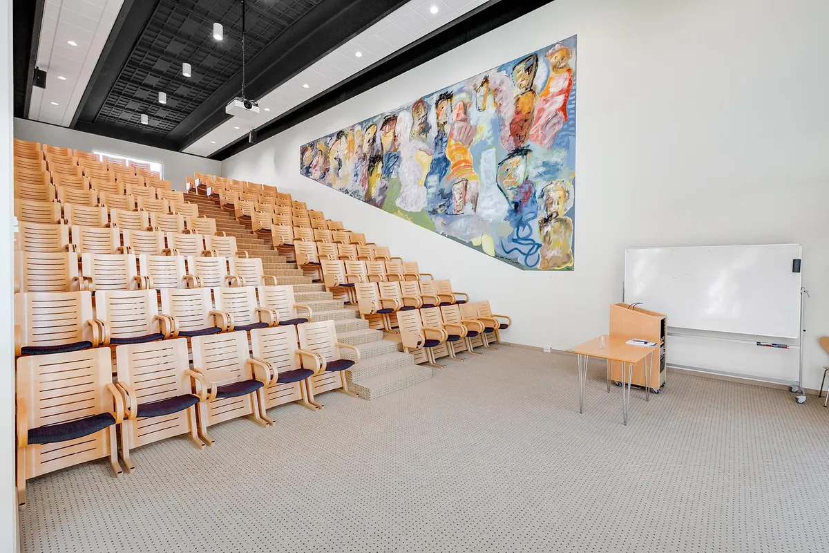 Auditorie med træstole og puder og et stort maleri på væggen og hvide tavle med en lille bord og en stol i hjørnet