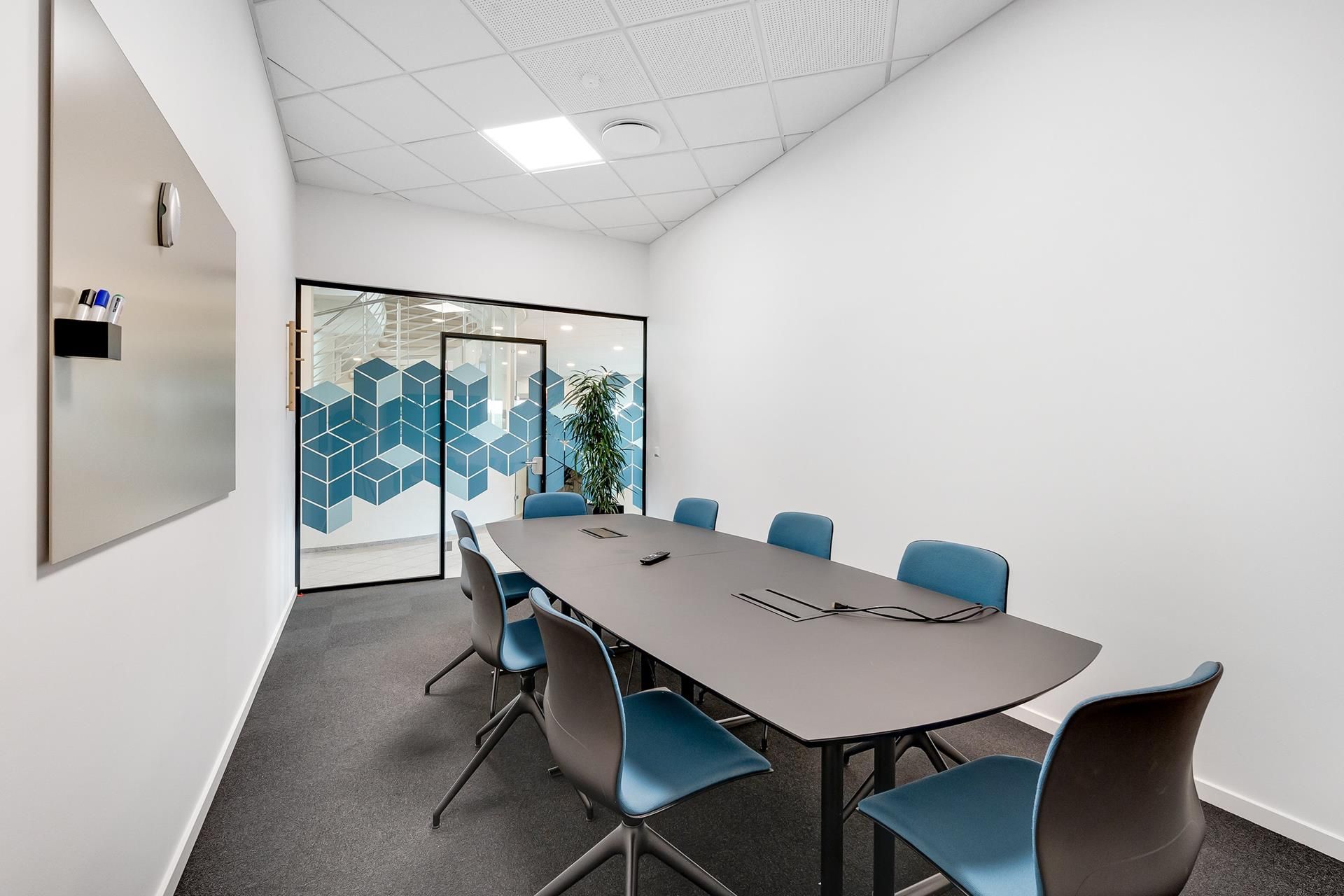 En mødesalon med moderne interiør, blå stole og et langt mødebord. Væggene er hvide, og der er et spejl med et skrivebord og