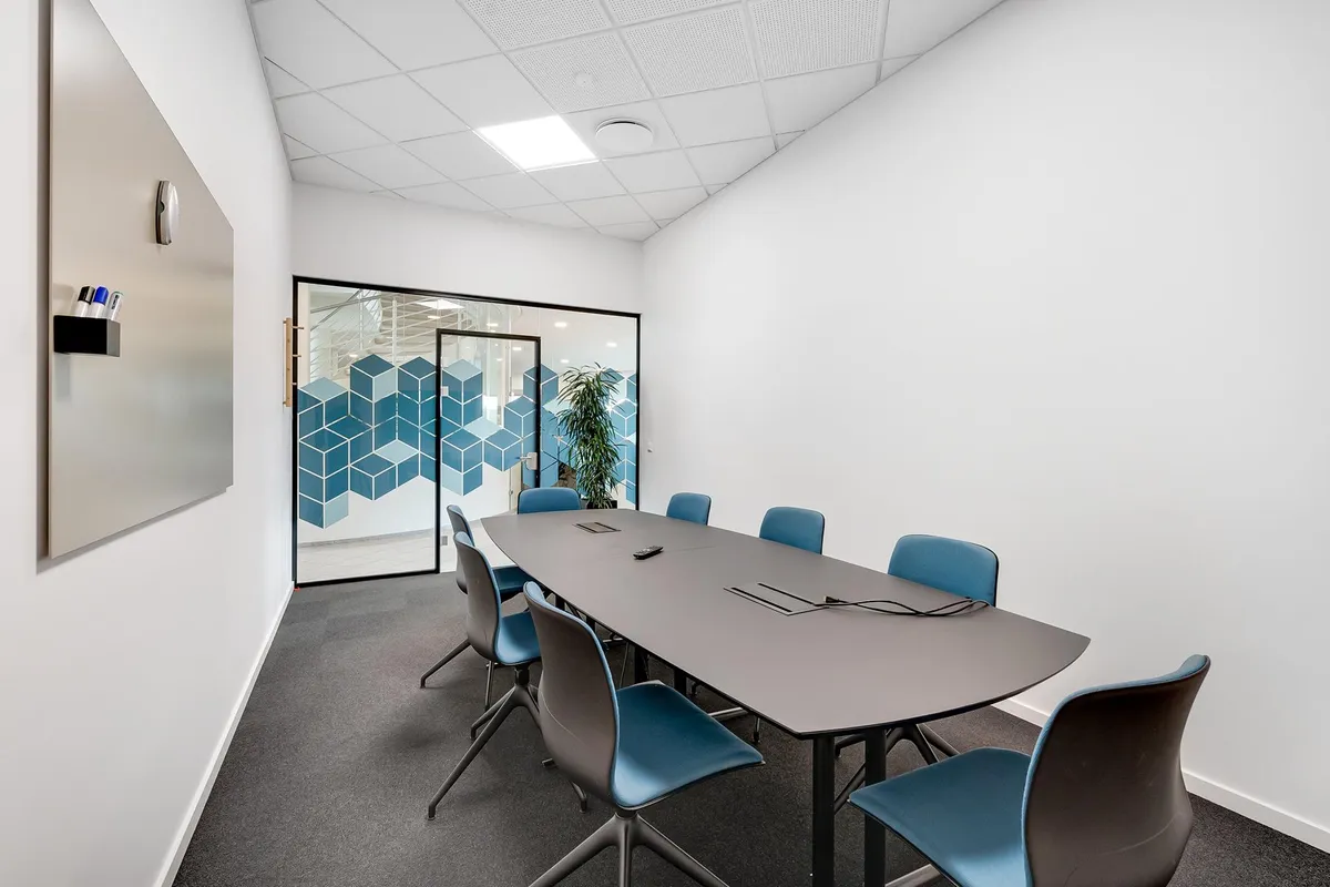 En mødesalon med moderne interiør, blå stole og et langt mødebord. Væggene er hvide, og der er et spejl med et skrivebord og