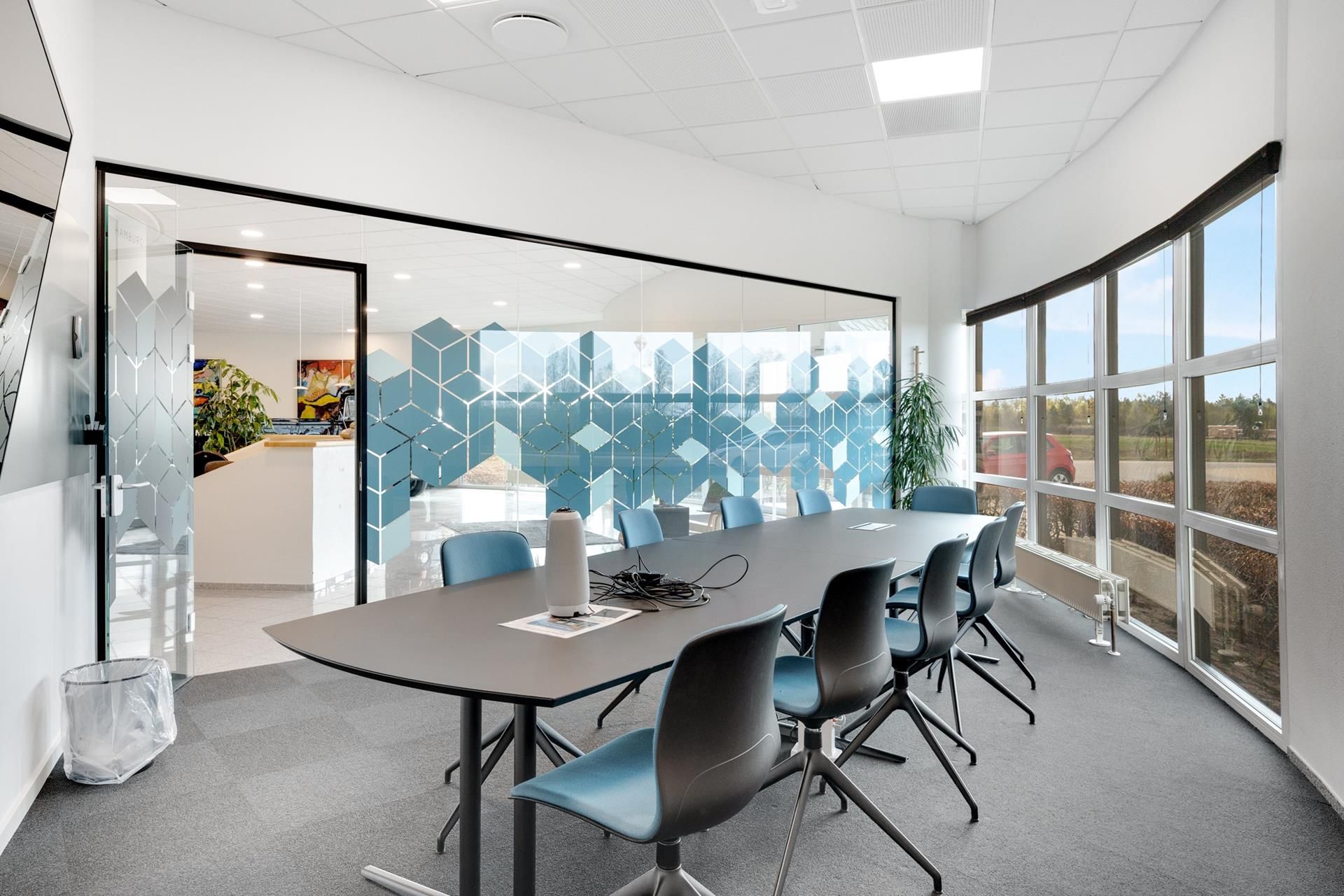 Kontor med runde vinduer og møbelgruppe med langt borde og stole. Væggen er hvid og gulvbelægningen er grå.