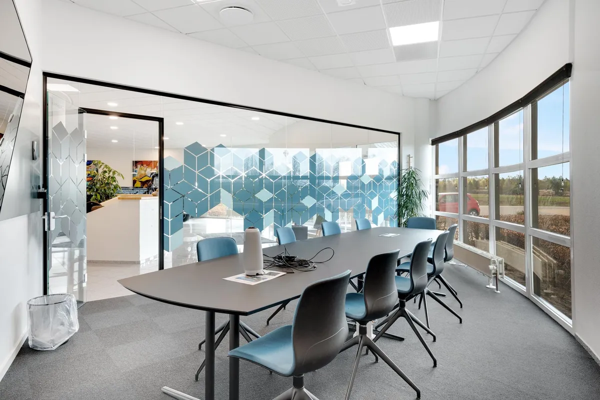 Kontor med runde vinduer og møbelgruppe med langt borde og stole. Væggen er hvid og gulvbelægningen er grå.