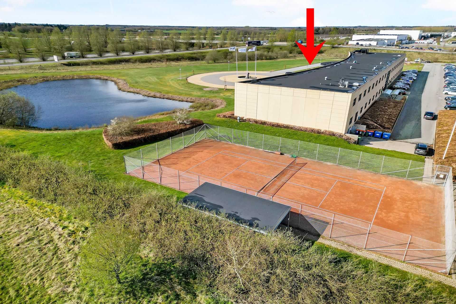 Tennisbaner med banehus og parkeringsplads. Et parkområde med vand og grønne arealer. En stor bygning med facade mod