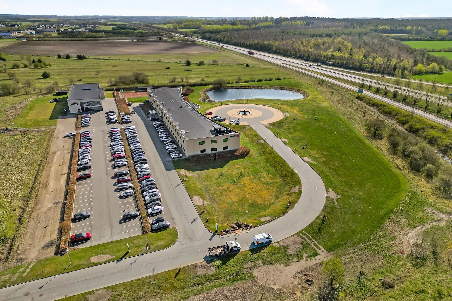 En bygning med parkeringsplads og motorvej i baggrunden, og en vandforsyning i forgrunden med grønne marker og buske på