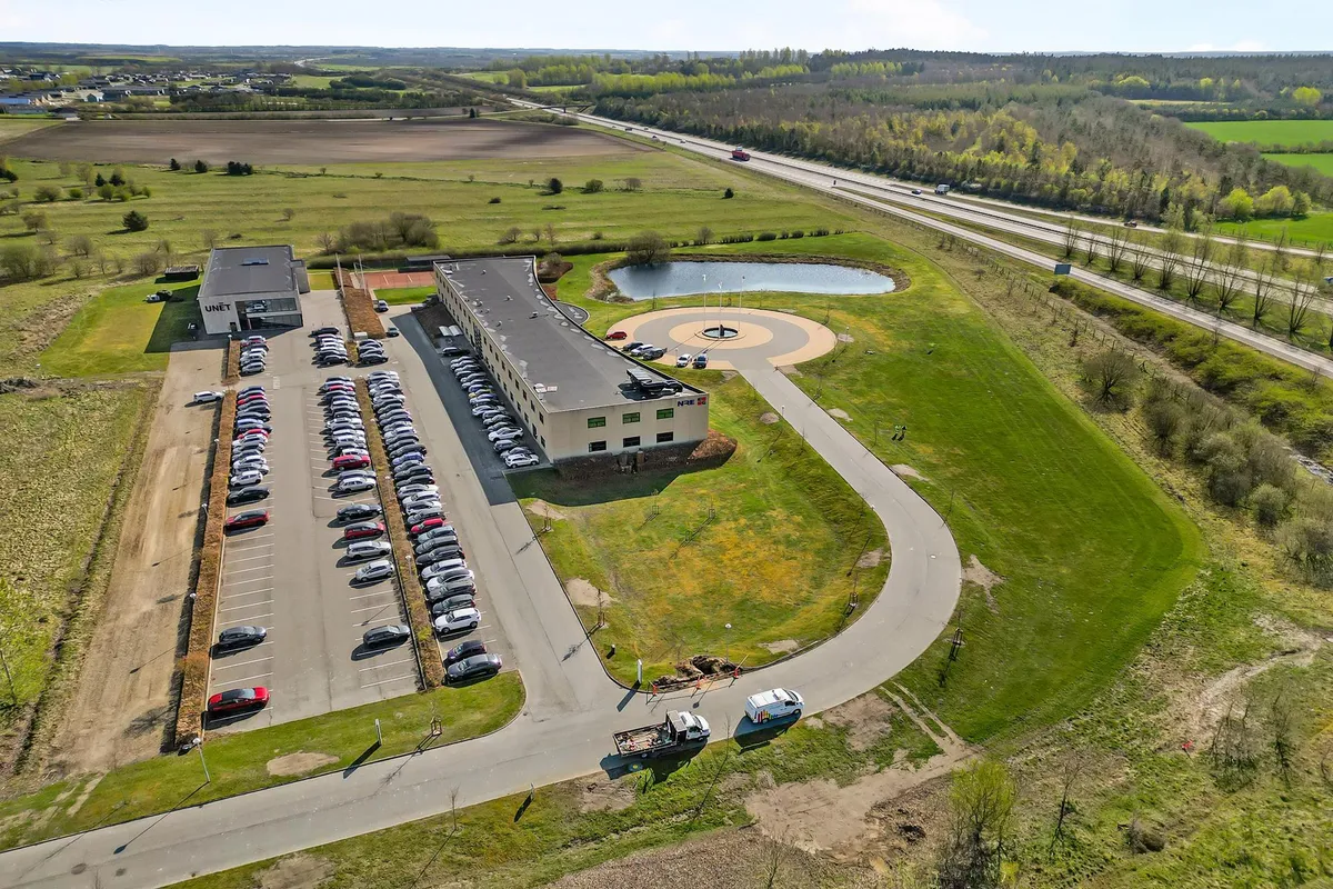 En bygning med parkeringsplads og motorvej i baggrunden, og en vandforsyning i forgrunden med grønne marker og buske på